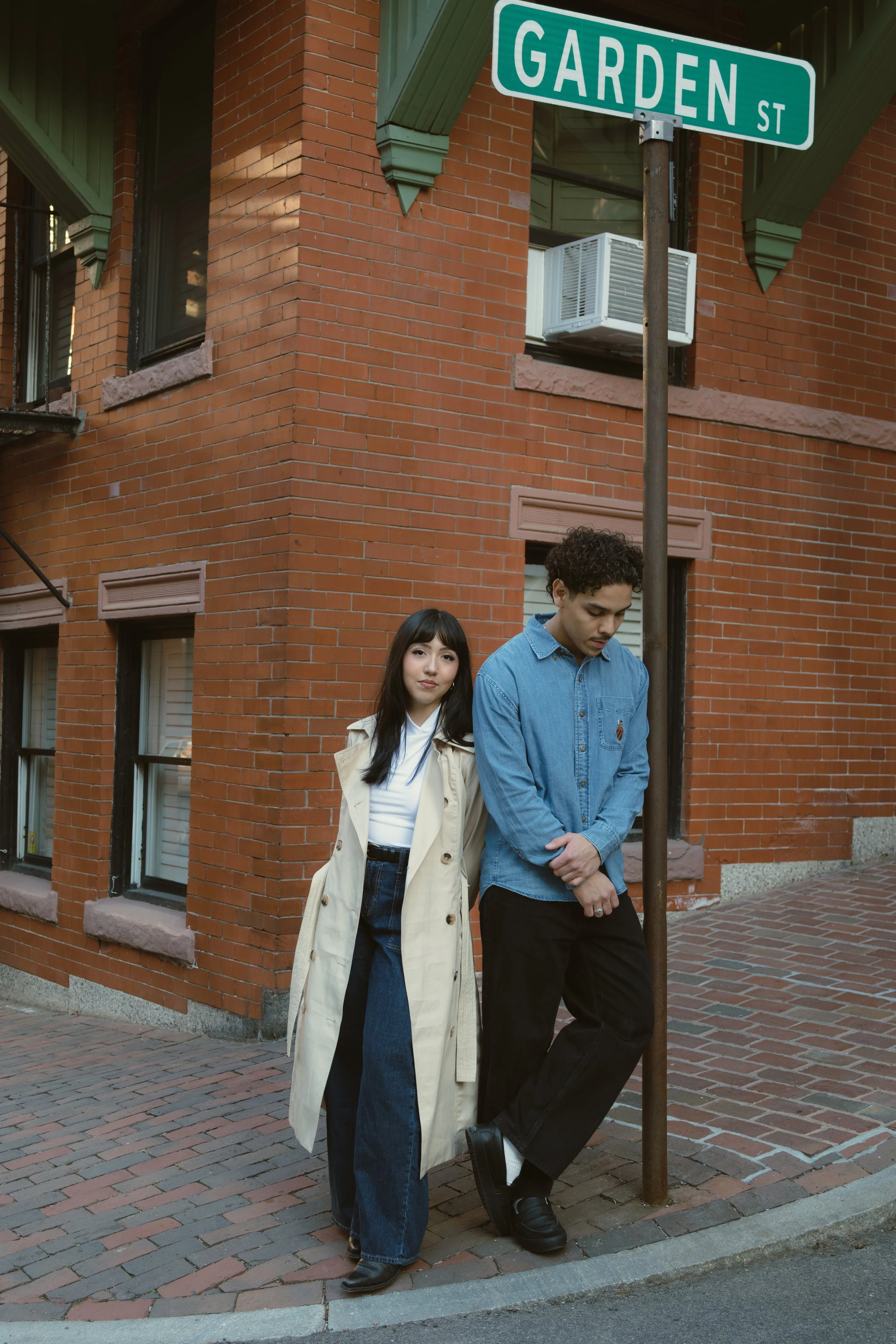 Two young people standing on a sidewalk near a brick building with a street sign that reads 'GARDEN ST.' The woman is wearing a beige trench coat and jeans, and the man is wearing a blue denim shirt and black pants, leaning against a pole.