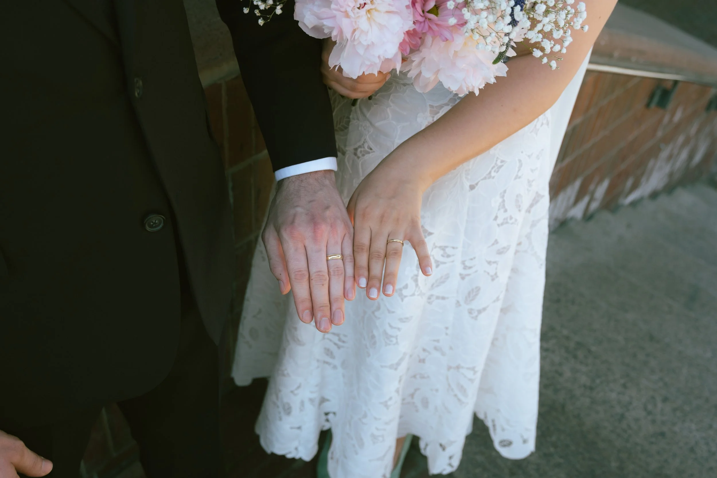 Close-up of a bride and groom holding hands, showing wedding rings, with the bride holding a bouquet of pink and white flowers, wearing a white lace dress, and the groom dressed in a black suit, outdoors against a brick wall.