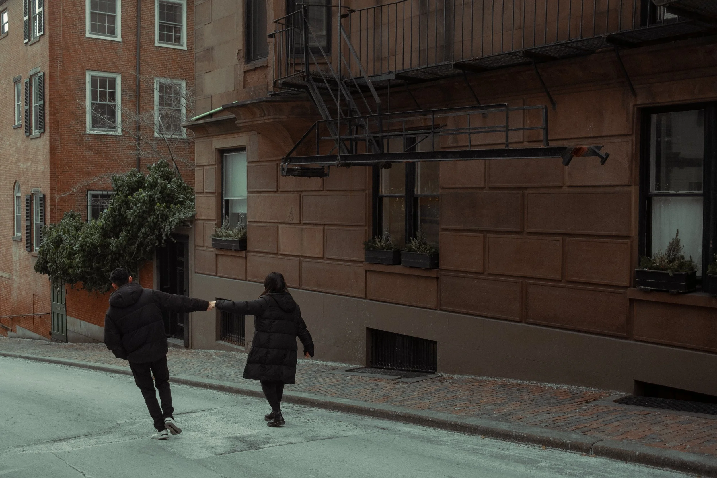 Two people walking hand in hand on a city street, holding hands behind their backs, near brick buildings and an alley.