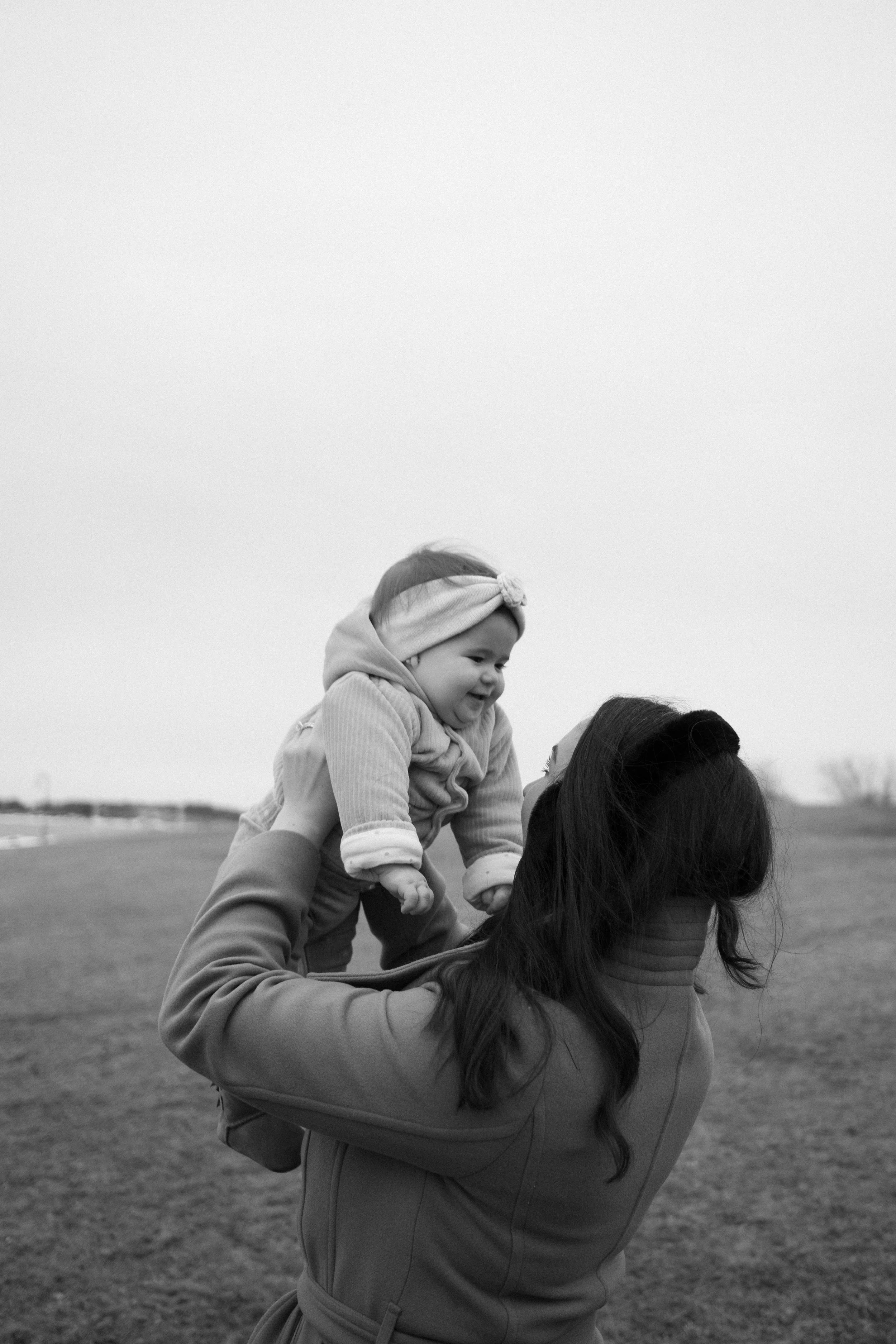 A woman with long hair wearing a jacket and headband lifting a smiling toddler girl in a hoodie and headband outdoors in a field, both enjoying a moment together.