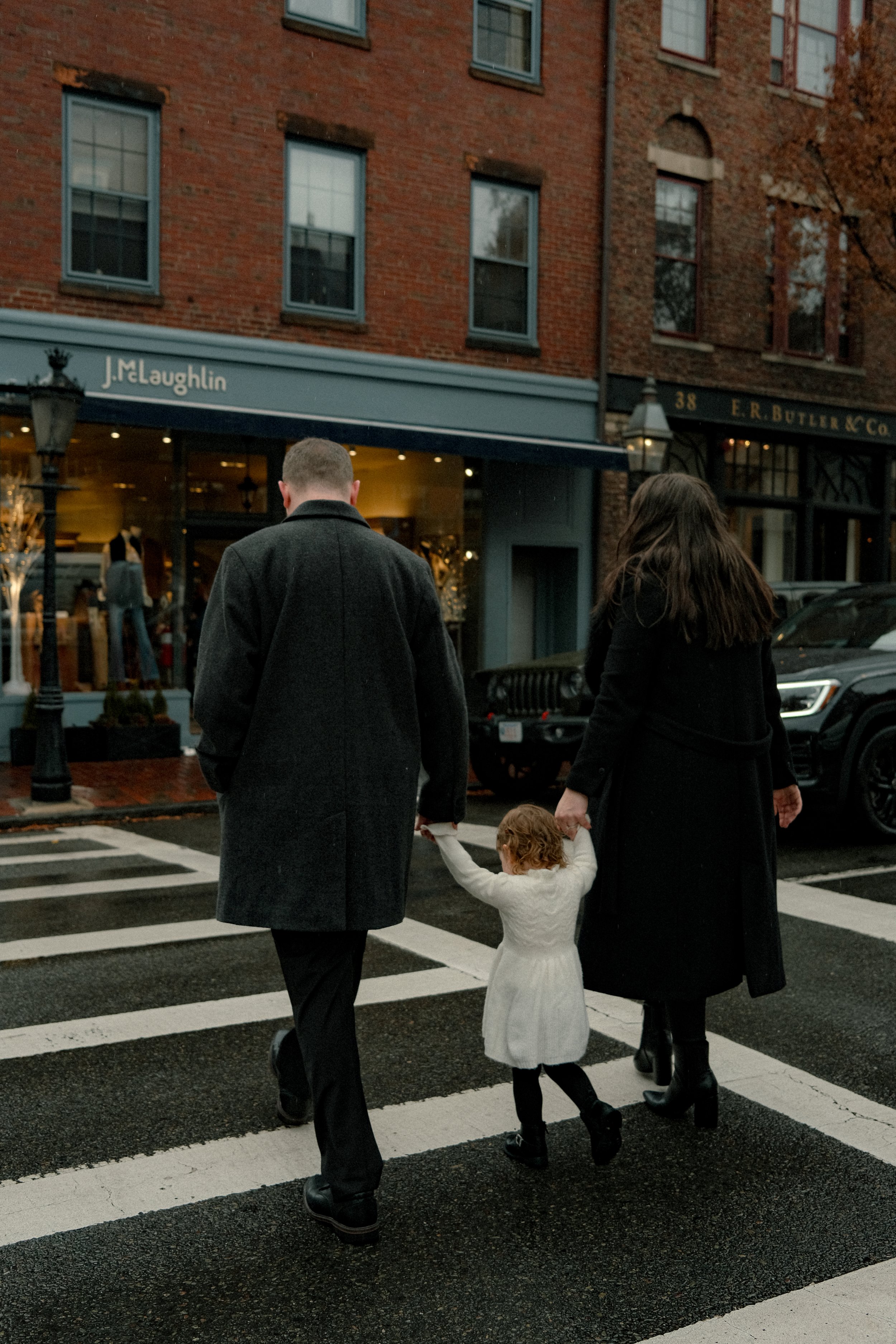 A family walking across a crosswalk on a rainy day in an urban area, holding hands. The father and mother are dressed in dark coats, and their young daughter is wearing a white dress and black tights.