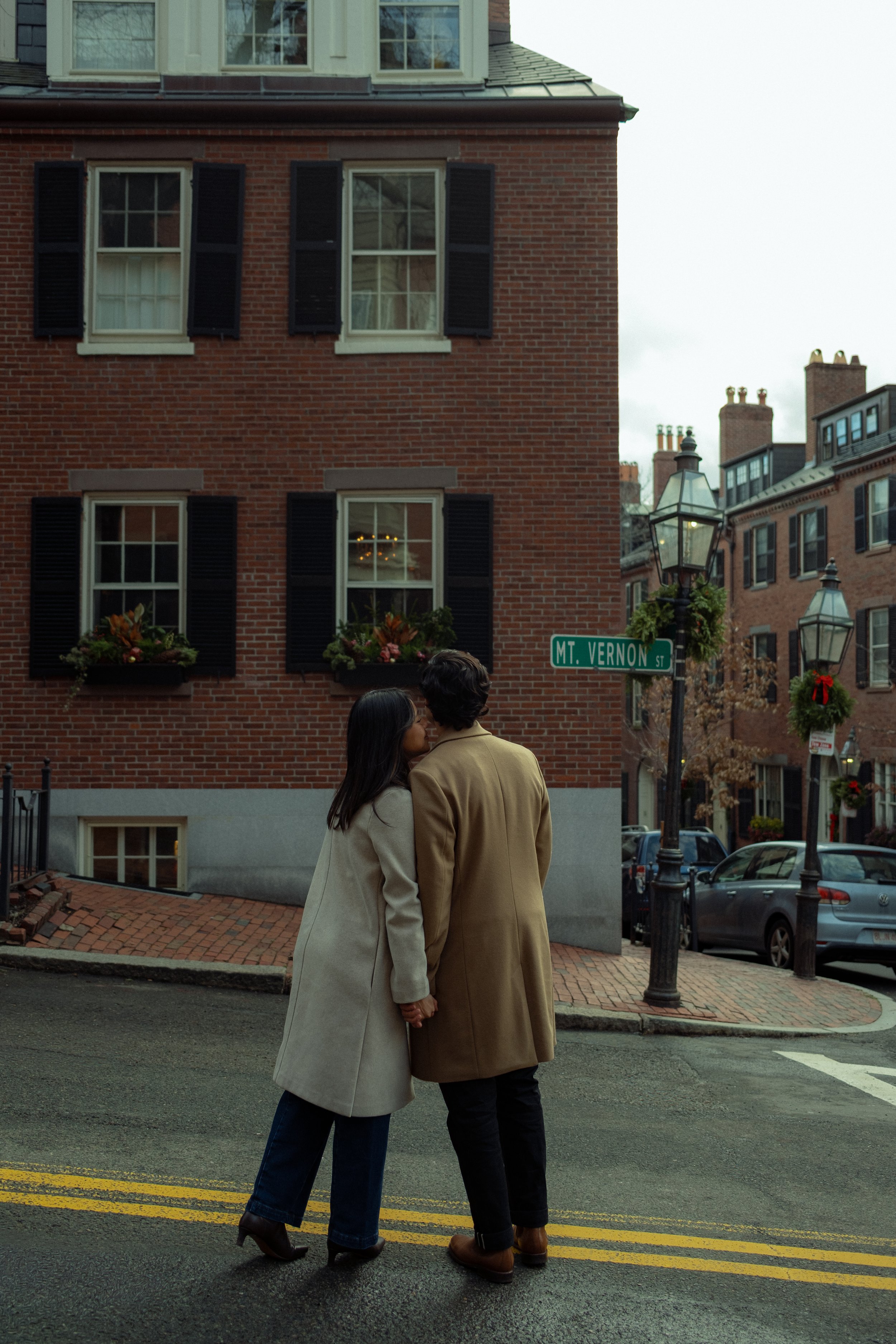 A couple kissing on a city street corner with a brick building, street signs, parking meters, and parked cars in the background during daytime.
