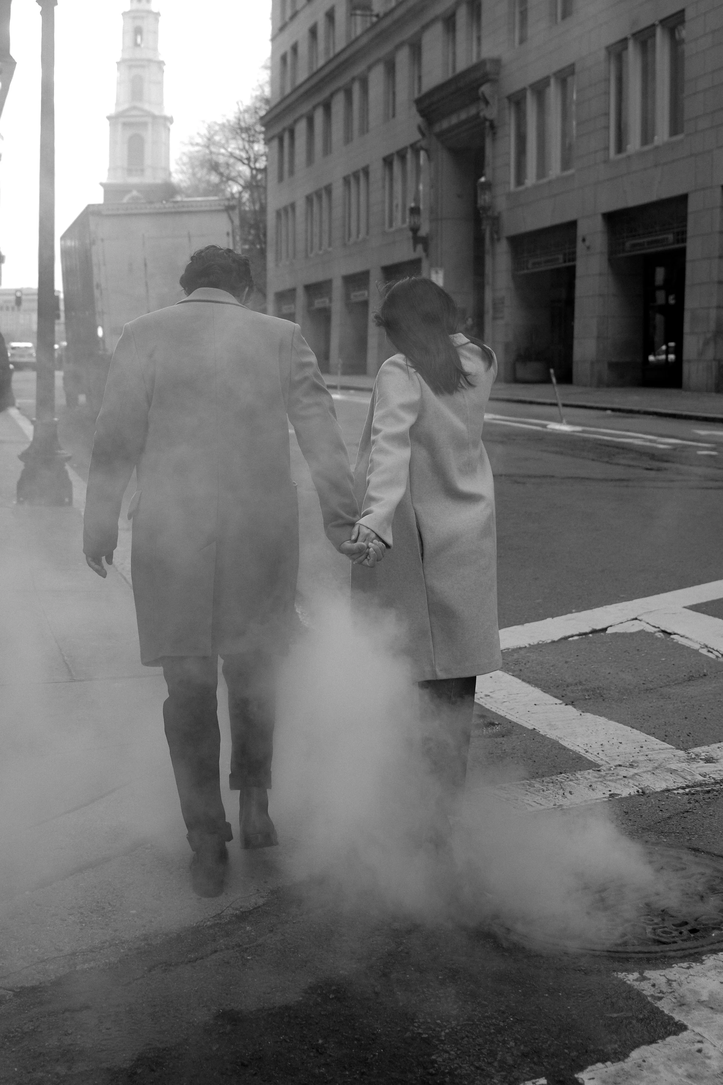 A black and white photo of a couple walking hand in hand on a city street, near a manhole cover with steam rising from it, surrounded by tall buildings and a street corner.