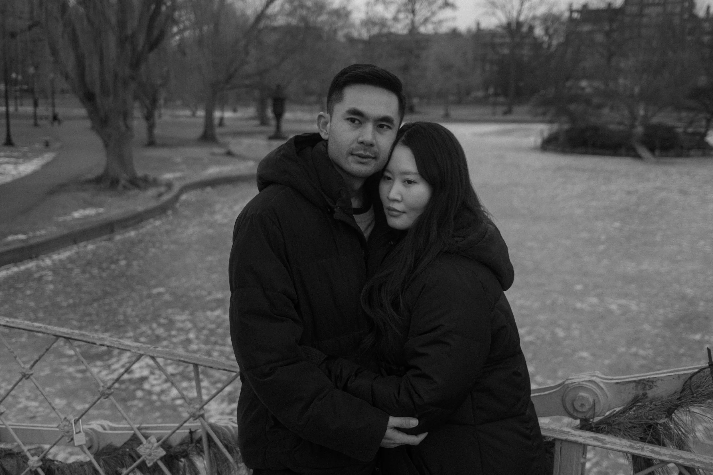 A black and white photo of a couple standing outdoors on a park bench, holding hands and embracing. The man has short dark hair and appears to be looking slightly to the left, while the woman has long dark hair and is looking to the right. There are trees and a walking path in the background, with some patches of snow on the ground.