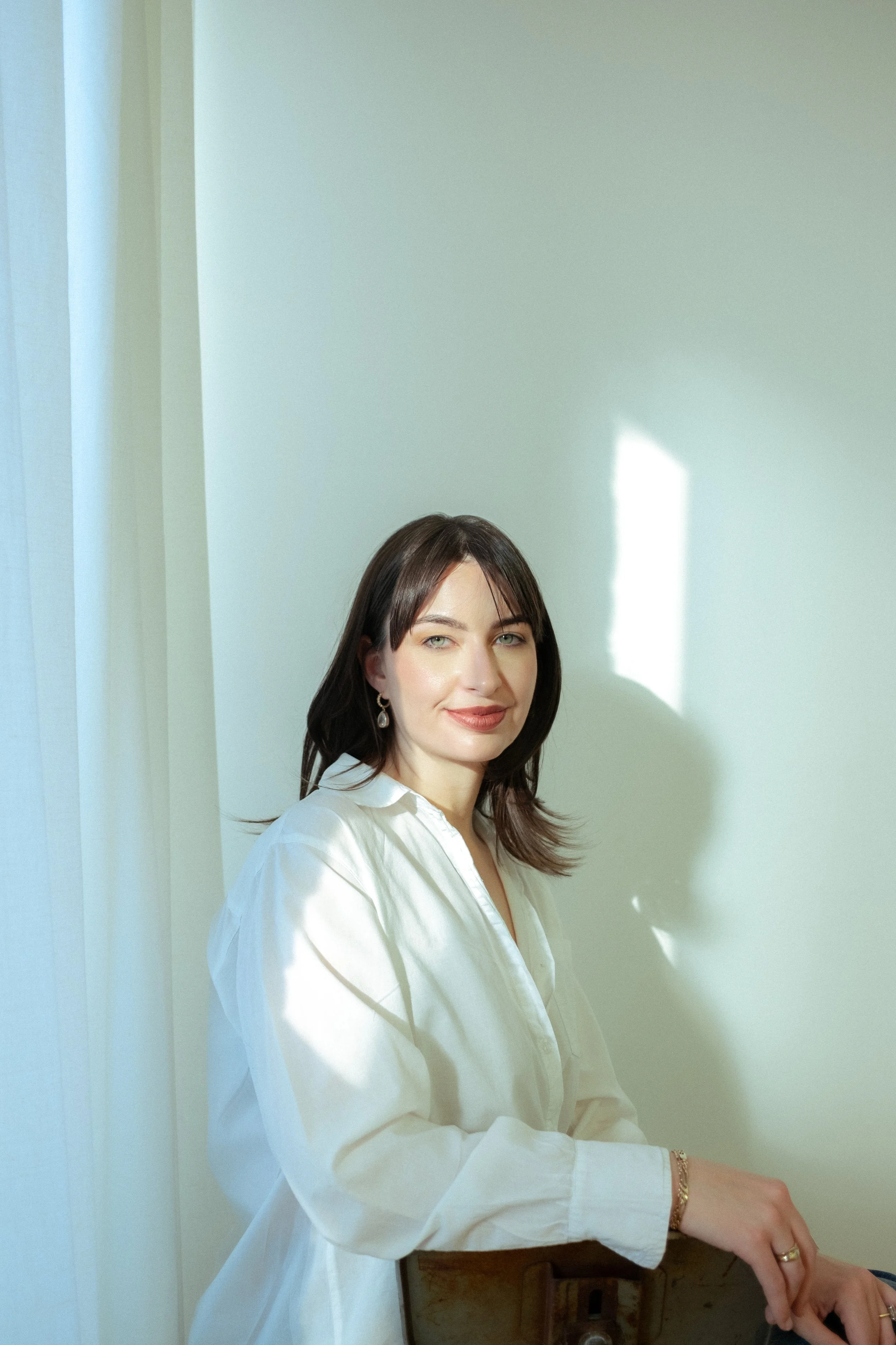 A woman with brown hair and blue eyes sits next to a white wall with light shining on her face, wearing a white shirt, earrings, and bracelets.