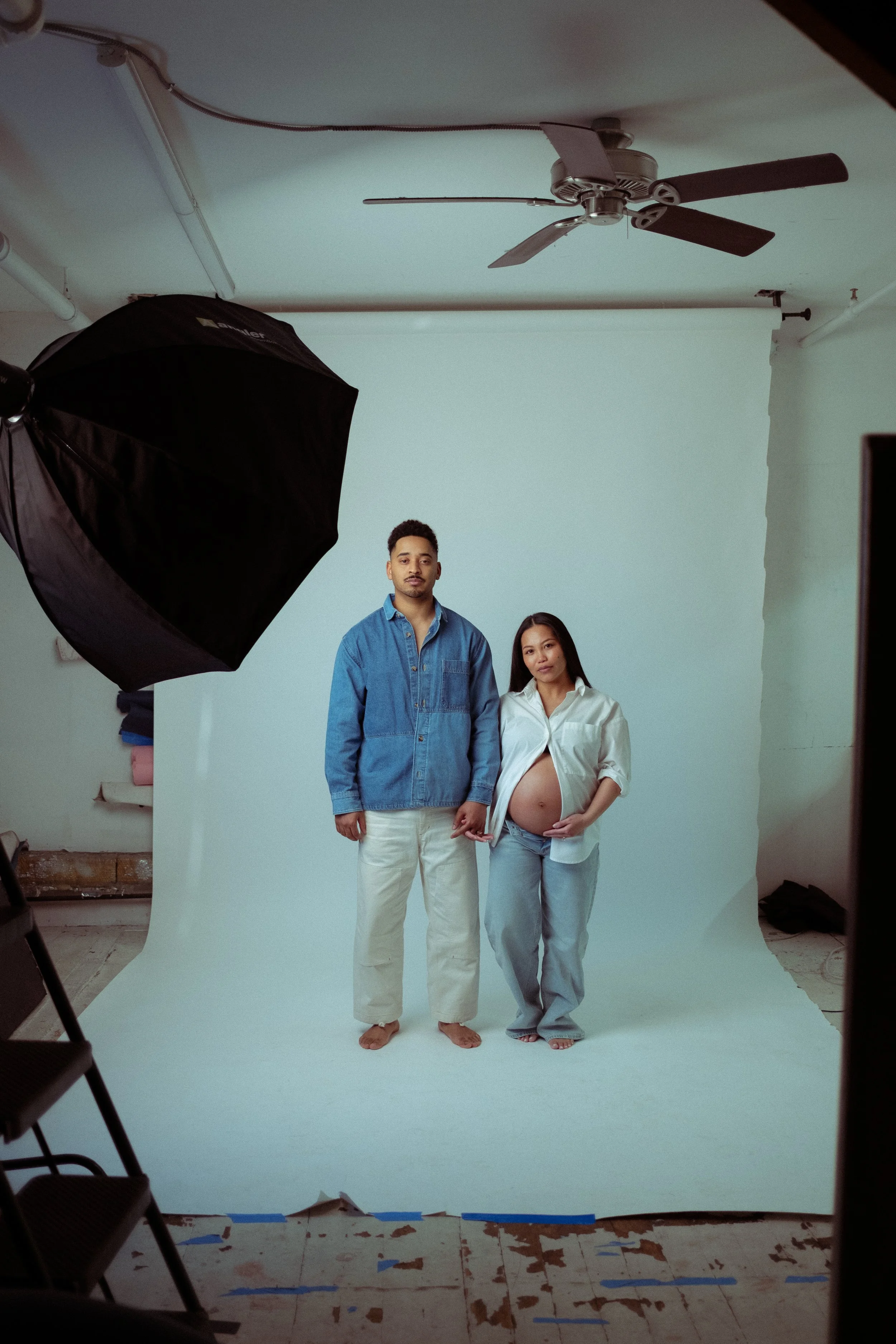 A pregnant woman and a man standing together on a white photo backdrop in a photography studio, with photography equipment visible around them.