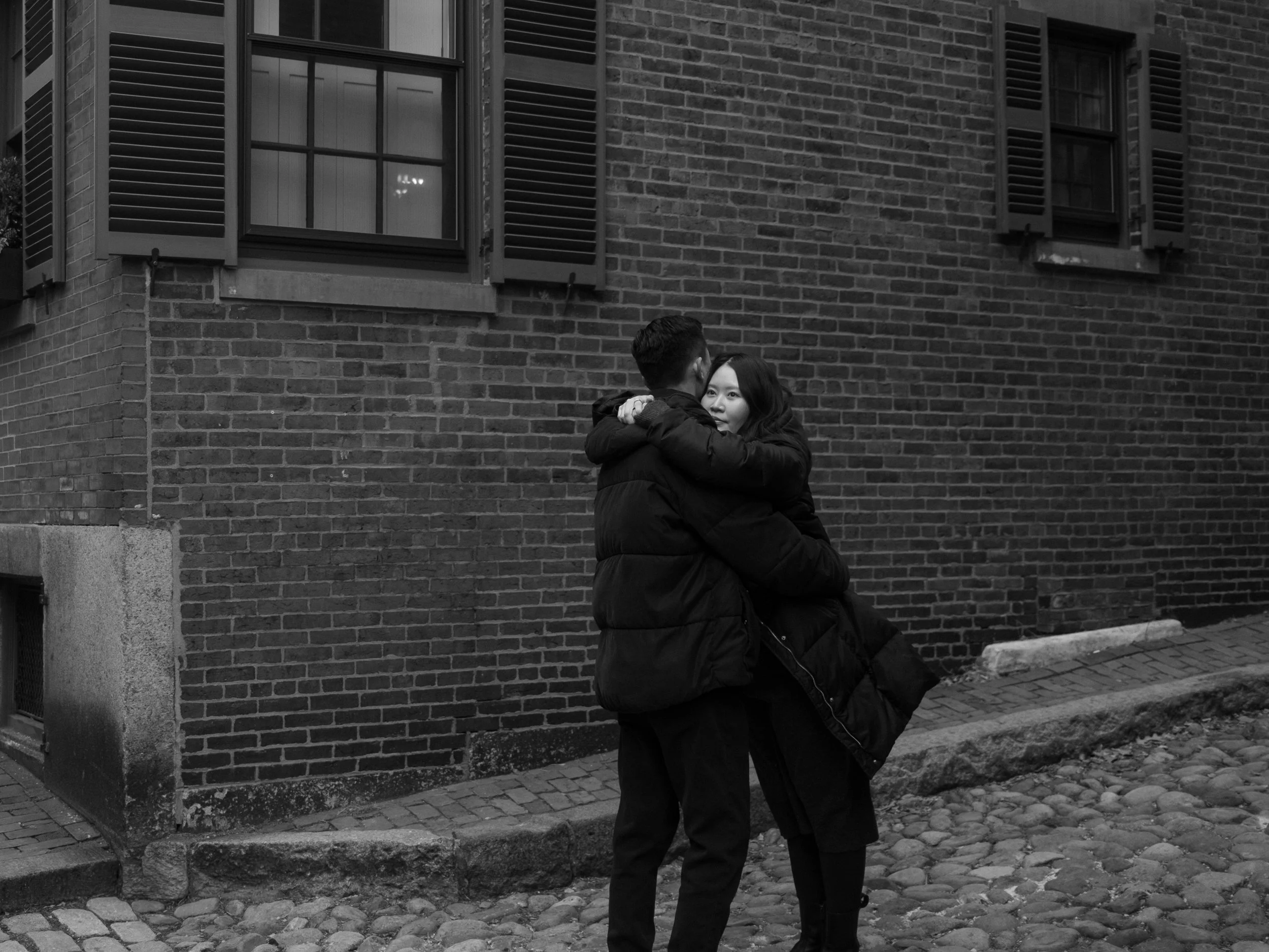 A man and woman embrace in a hug on a cobblestone street in front of a brick building with several windows.