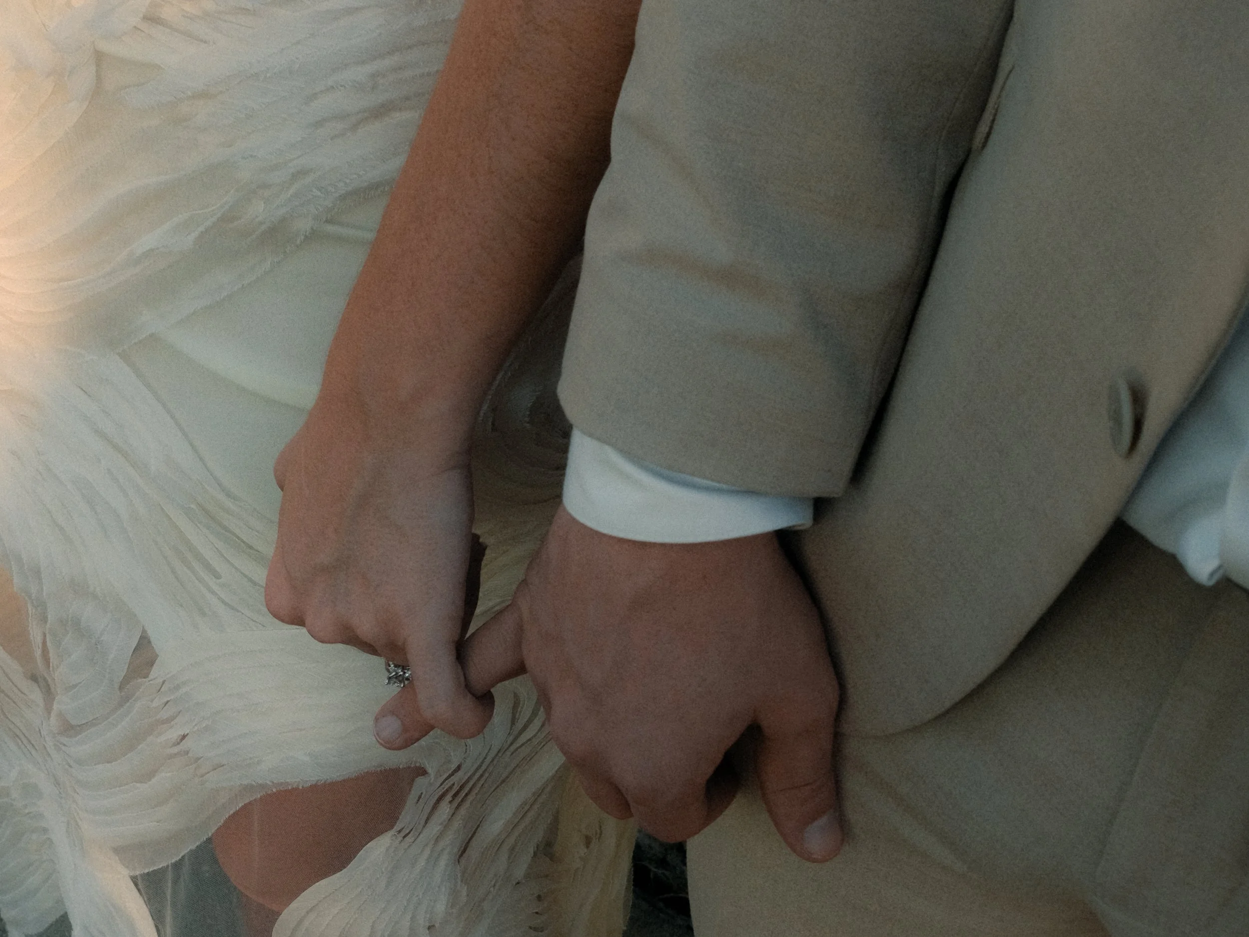 Close-up of two hands holding each other, one hand appears to be female with rings, the other male, with a beige jacket and white shirt sleeve visible.