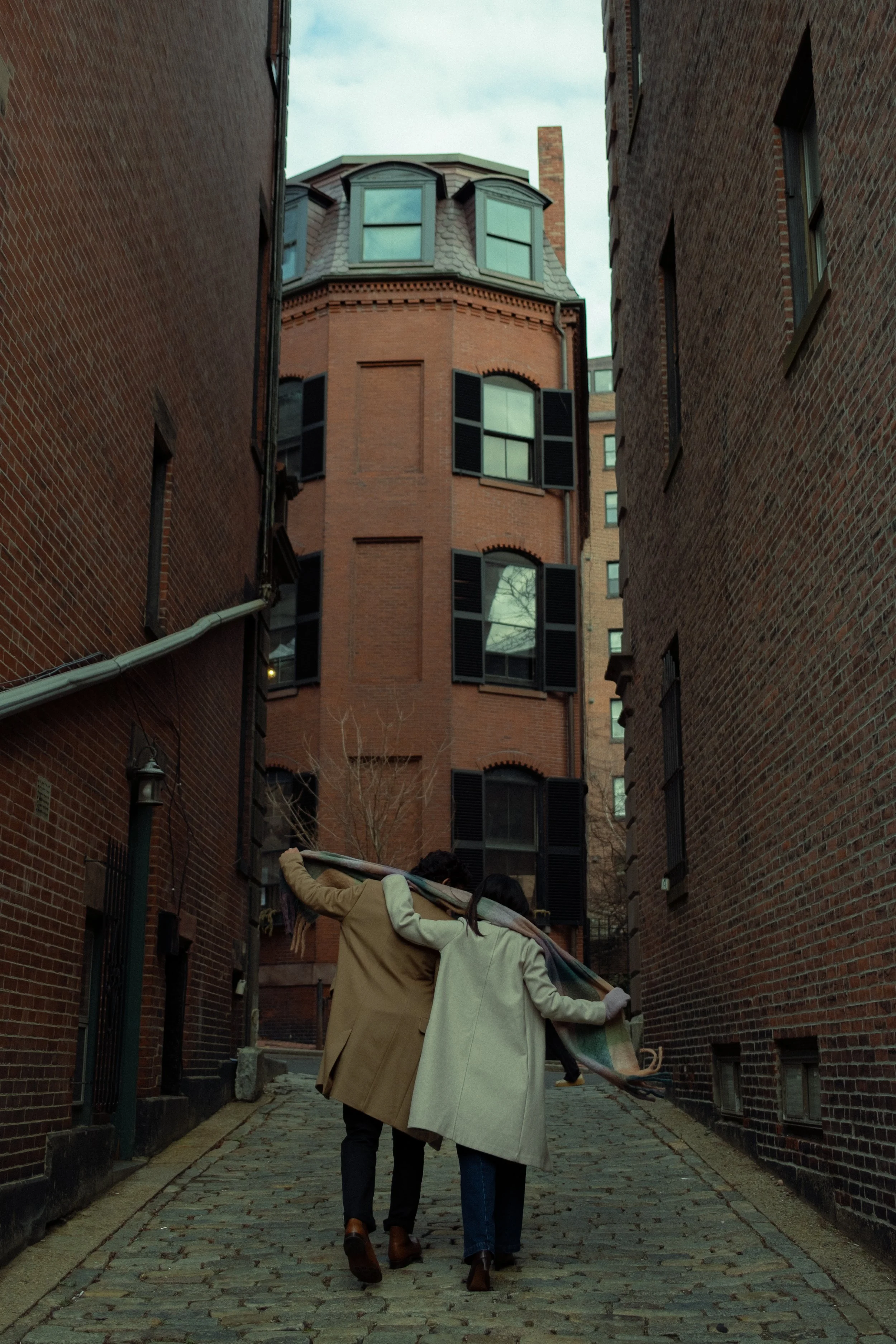 Two women walking down a cobblestone alleyway between tall brick buildings, carrying a rolled-up carpet on their shoulders.