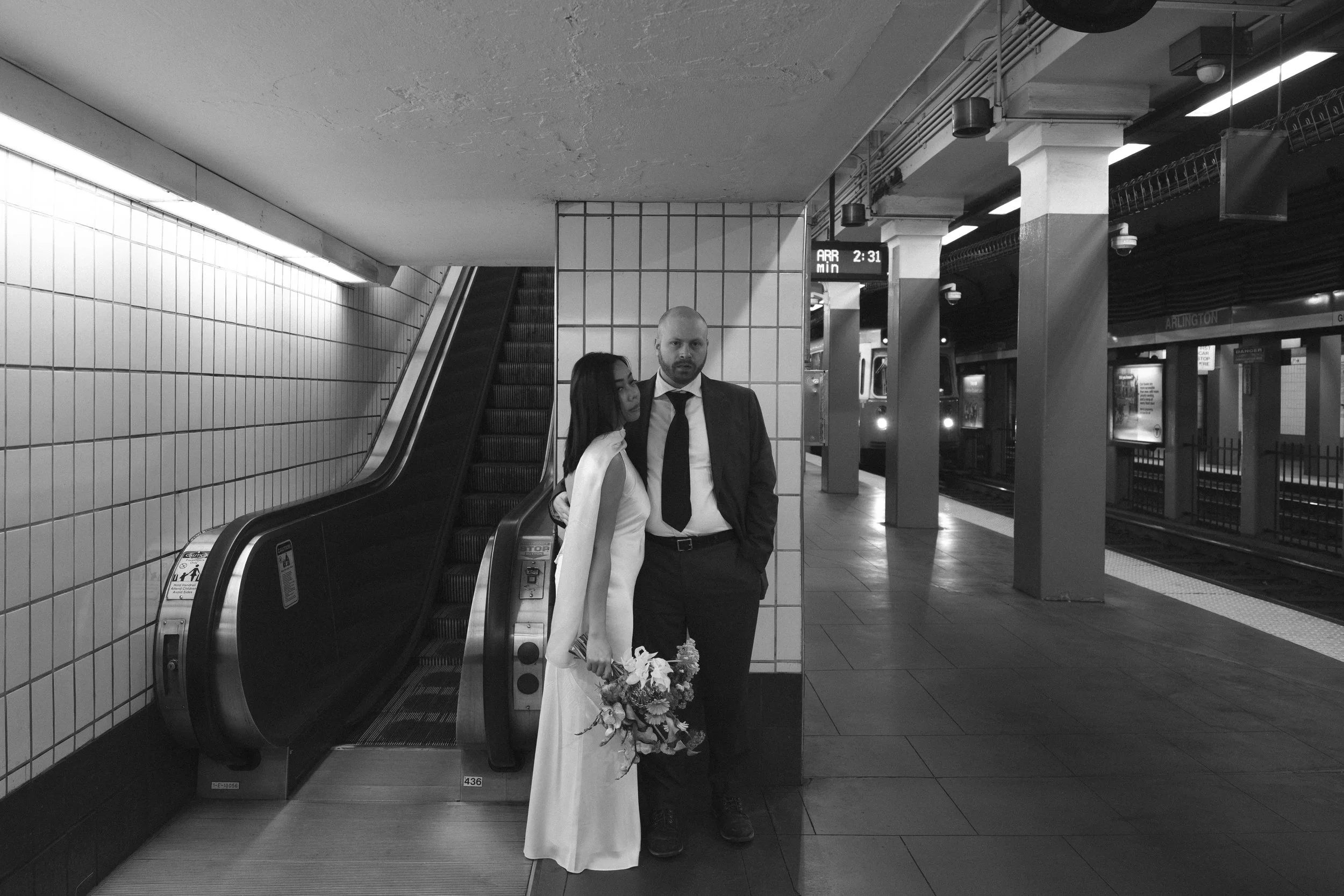 A couple, the woman in a white dress holding a bouquet of flowers and the man in a suit, standing on an escalator at a subway station.