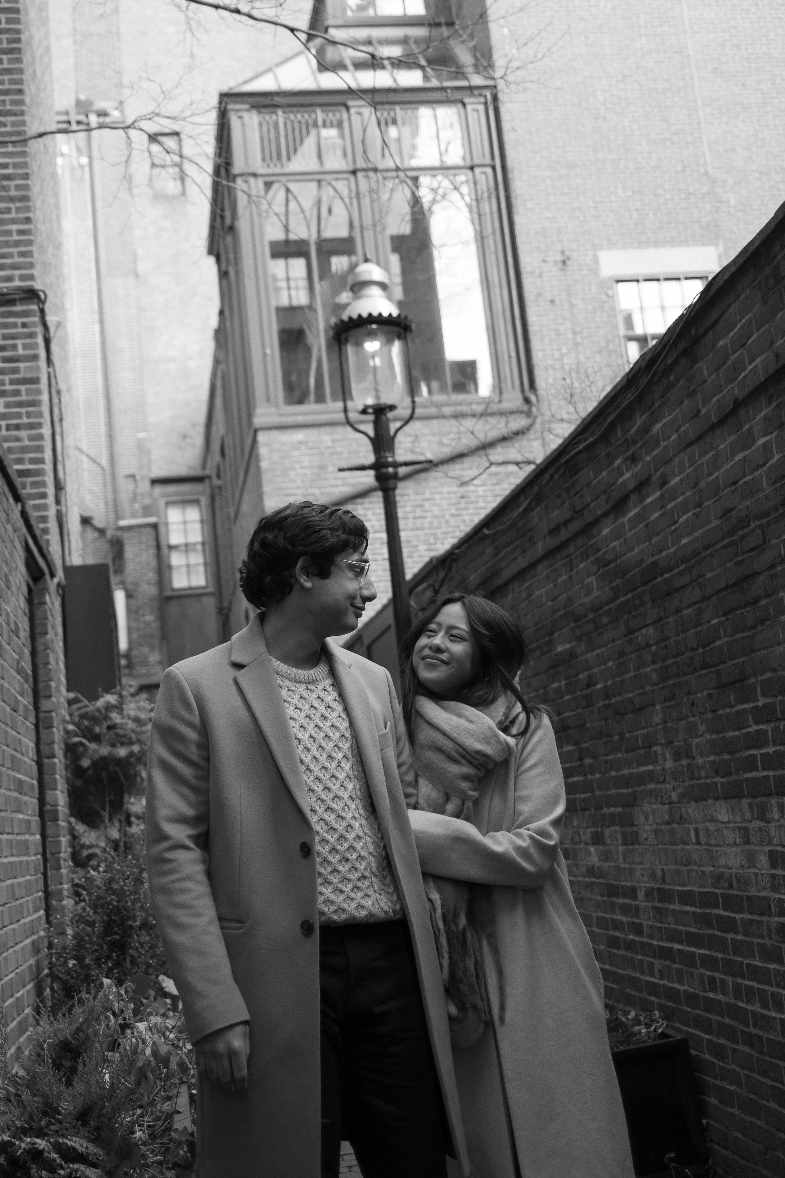A black and white photo of a man and woman standing together in a narrow alleyway with brick walls on both sides. The man is wearing glasses and a long coat, while the woman is smiling and embracing him, wearing a scarf and coat. A street lamp and tall building with bay windows are visible in the background.