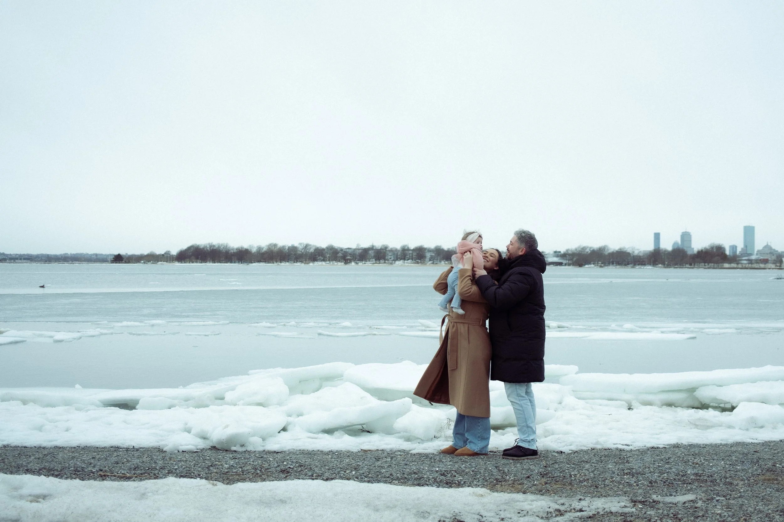 A family of three, a woman, a man, and a young child, standing by a frozen body of water during winter, embracing each other and smiling.