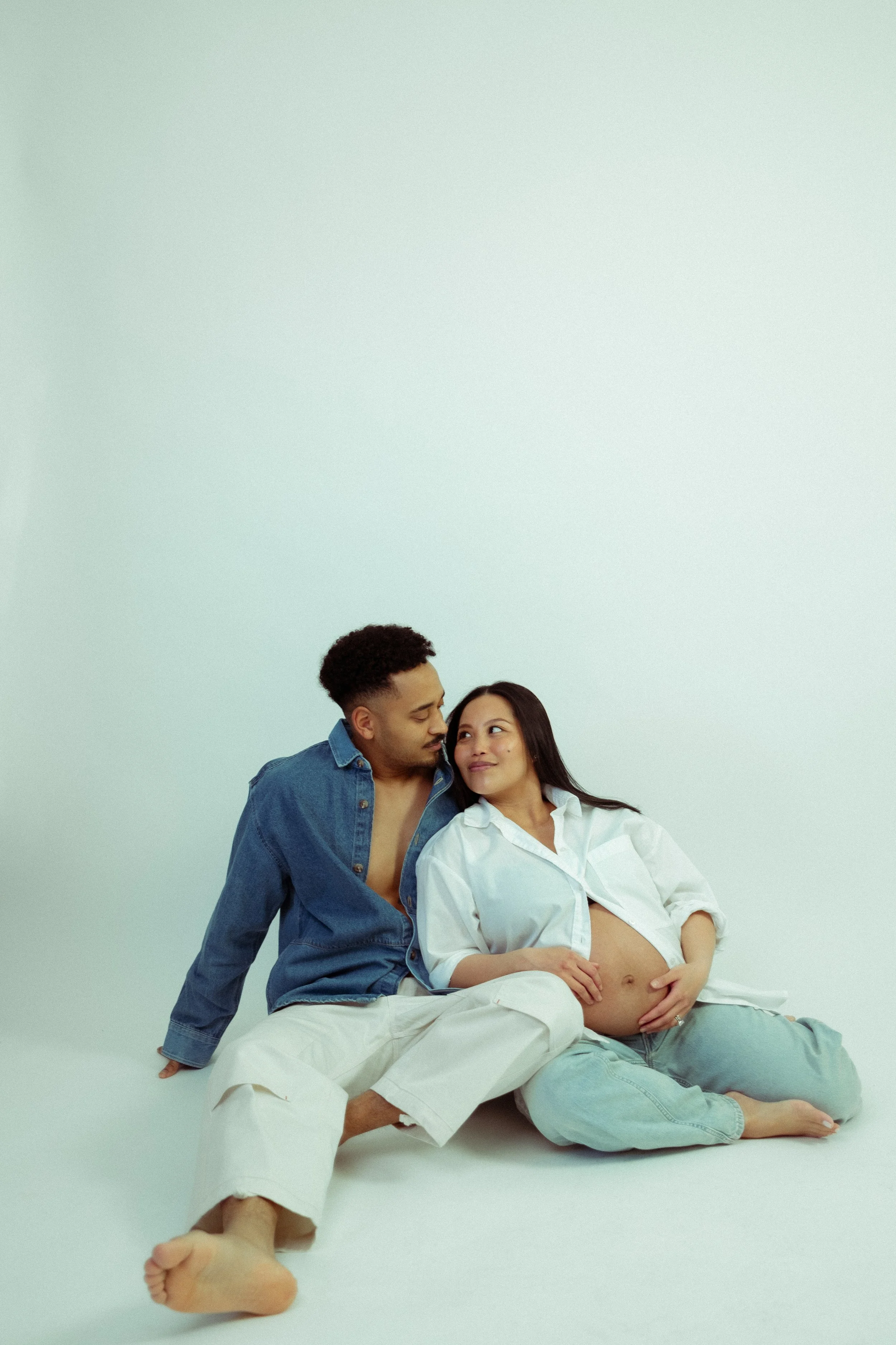 A pregnant woman and a man sitting on the floor close to each other, the woman is touching her belly and smiling while the man looks at her lovingly against a plain, light-colored background.