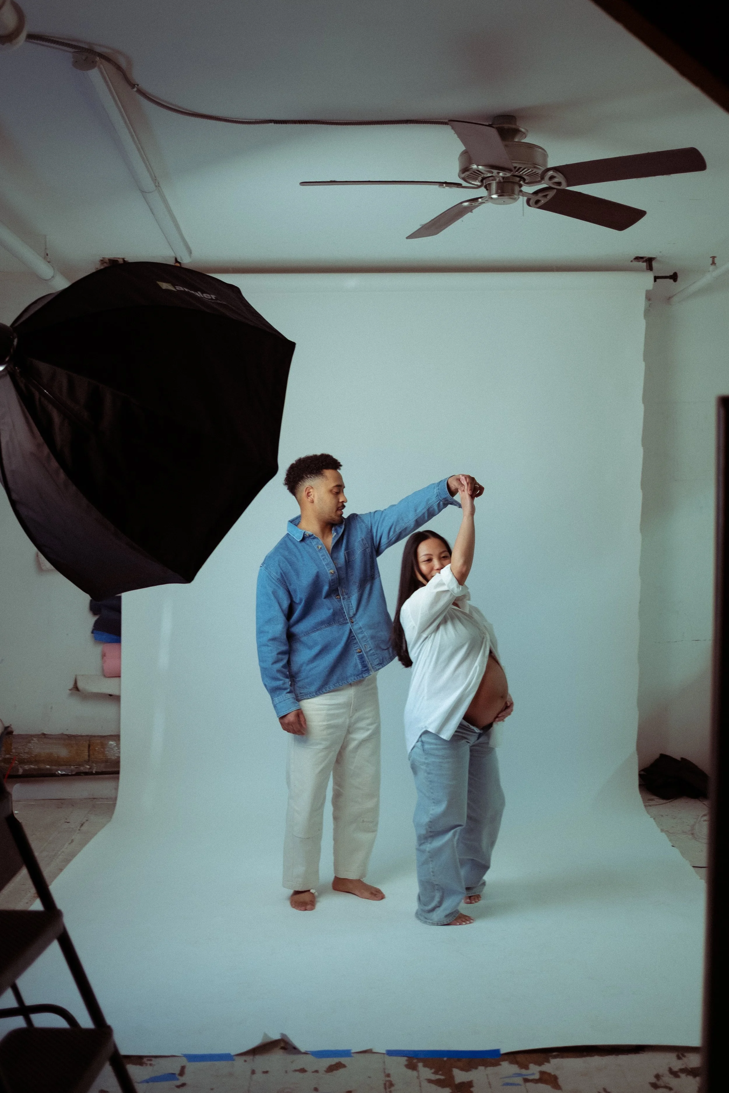 A pregnant woman and a man in a photography studio with a white backdrop. The woman is lifting her arm, and the man is holding her hand. Studio equipment, including a softbox light and ceiling fan, are visible.