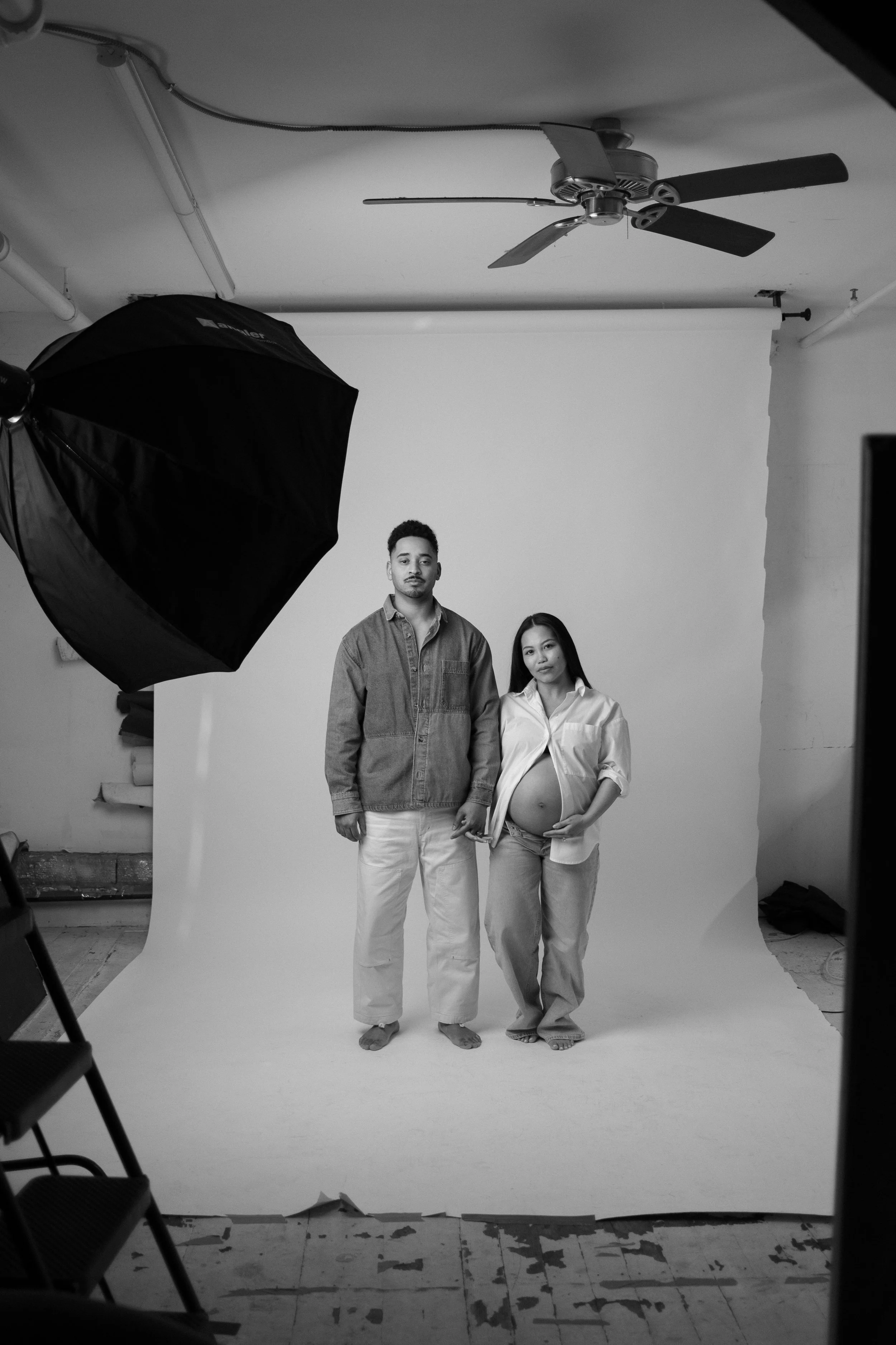 A black and white photo of two models standing in a photography studio on a seamless paper backdrop. The male model is wearing a shirt and white pants, and the female model is wearing a loose shirt, partly unbuttoned, revealing her pregnant belly, and light-colored pants. There is a large studio light with an umbrella to the left and the ceiling fan is visible above. Decorations on the floor indicate a worn floor surface.