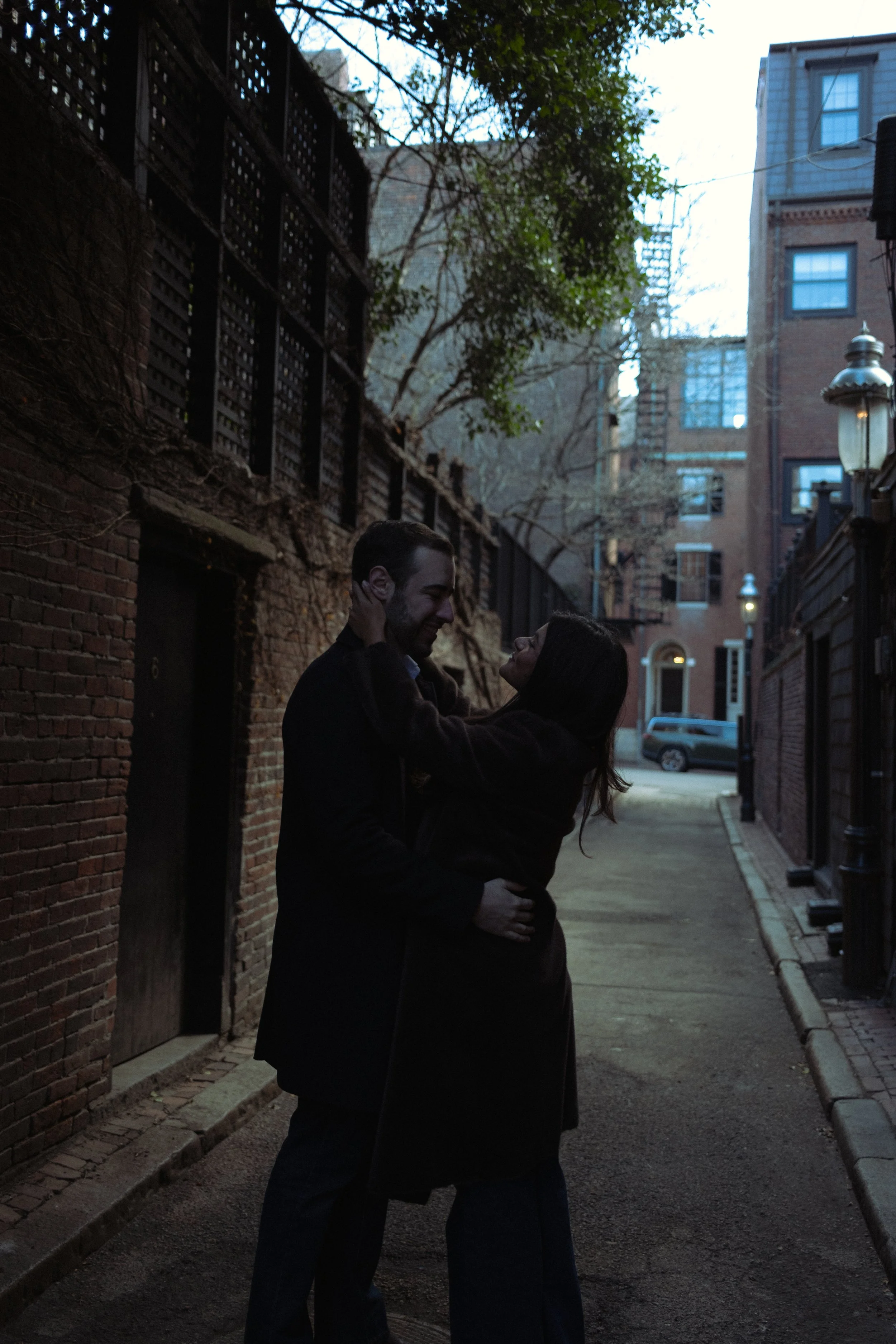 A man and woman standing close together in a dark, narrow alleyway, holding each other and smiling, with city buildings in the background.