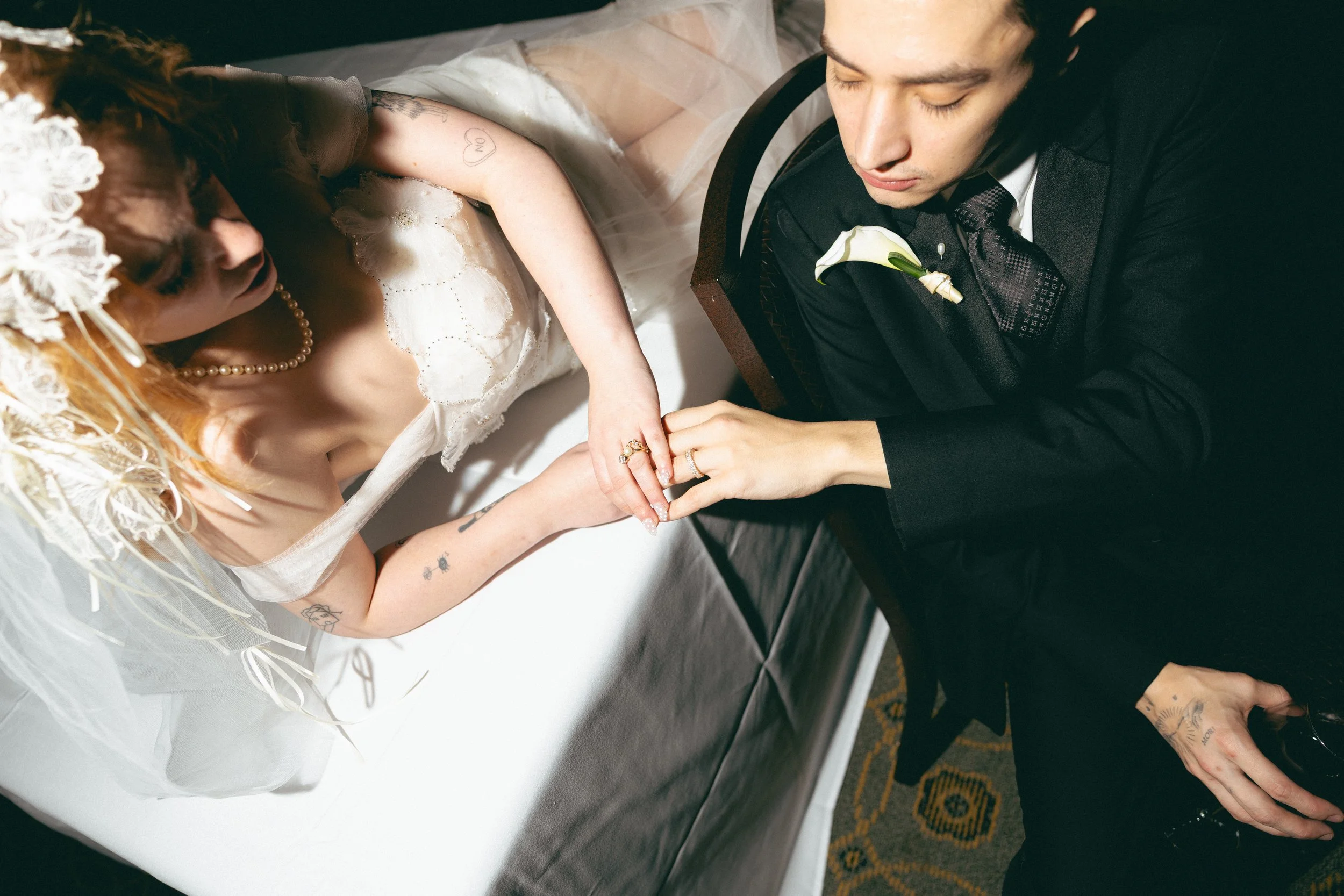 A newlywed couple holds hands during their wedding ceremony, with the bride dressed in a white lace gown and veil, and the groom in a black tuxedo with a white boutonniere, both with their eyes closed.