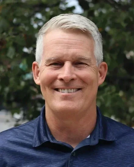A smiling man with gray hair wearing a blue suit jacket and light blue shirt, standing outdoors with green foliage in the background.
