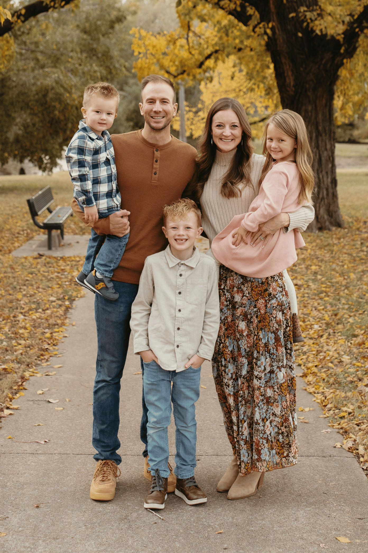 A family of five standing on a park sidewalk in autumn, with golden leaves on trees and ground, smiling at the camera.