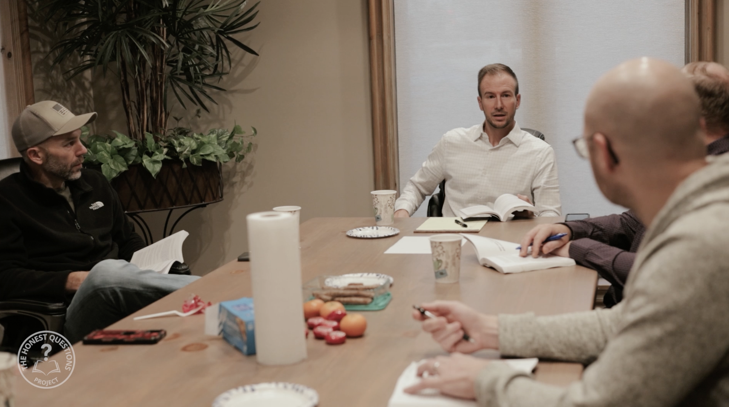A group of five men sitting around a conference table in a meeting room, with one man speaking and the others listening. The table has snacks, papers, and cups on it, and there are plants and a window in the background.