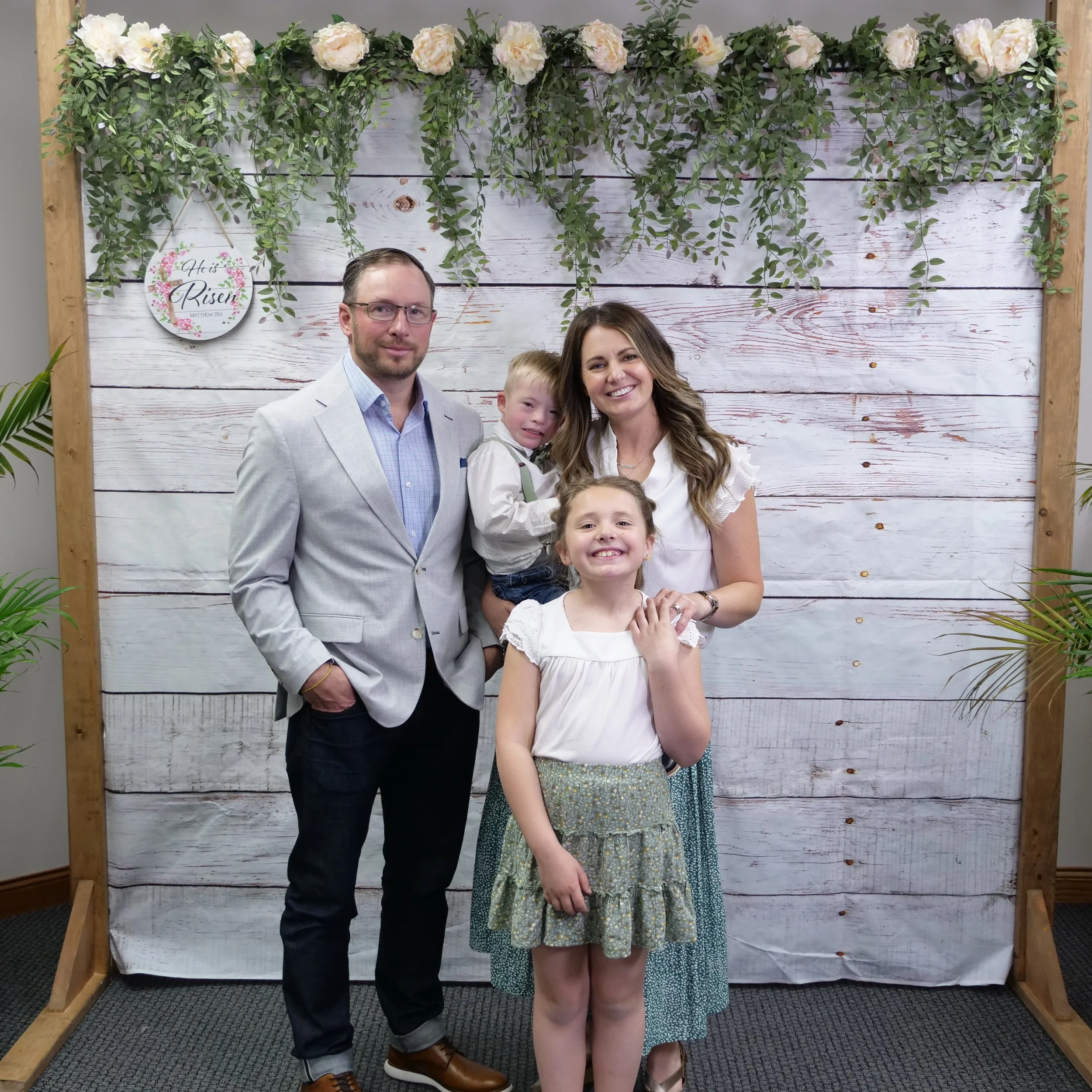 A family of four, including a man, woman, and two children, standing in front of a decorated backdrop with flowers and greenery at a celebration event.