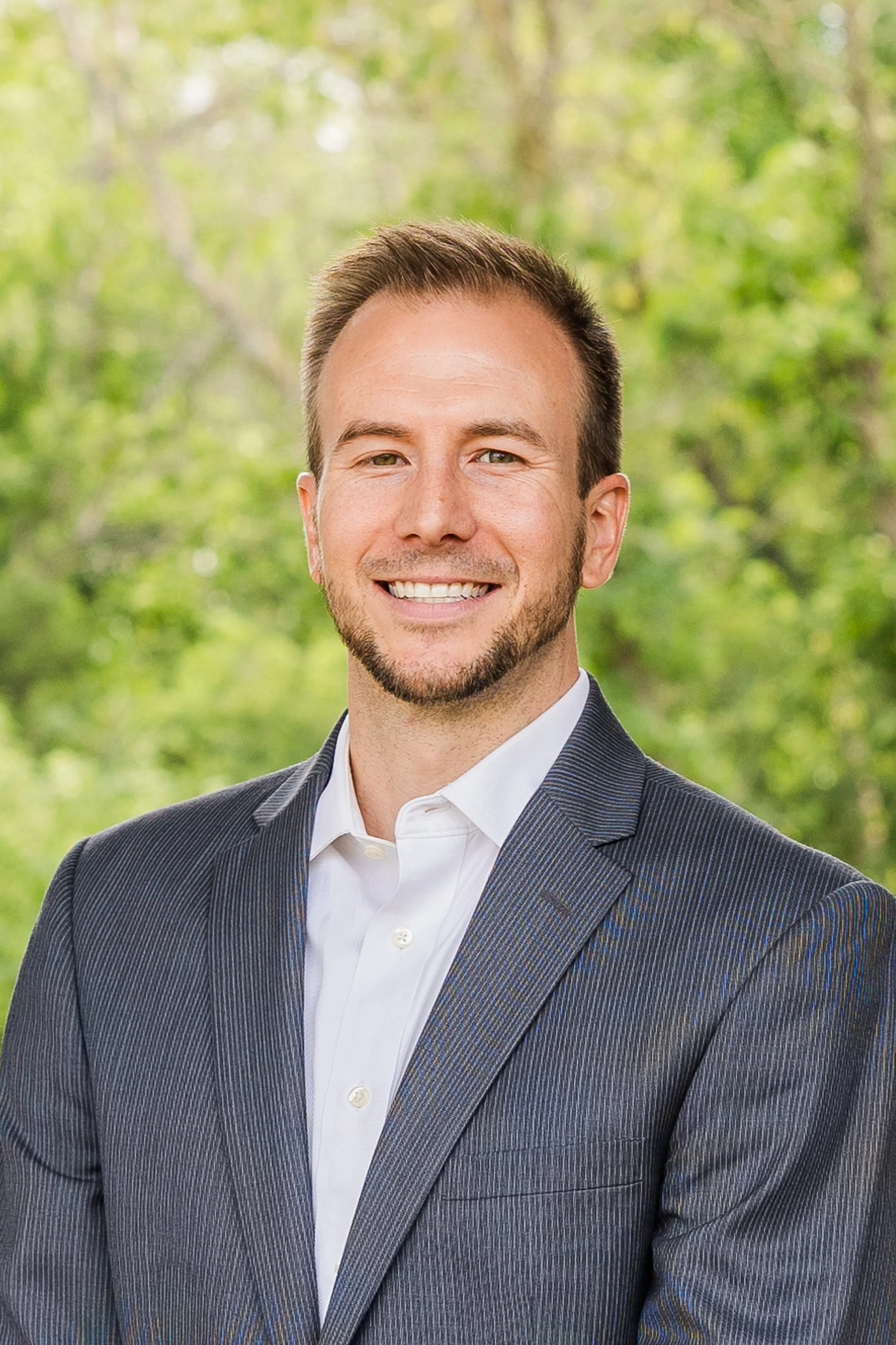 A smiling man in a gray suit and white shirt standing outdoors with green trees in the background.