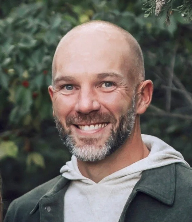 A smiling man with a beard and bald head outdoors, wearing a hoodie and jacket, surrounded by greenery.