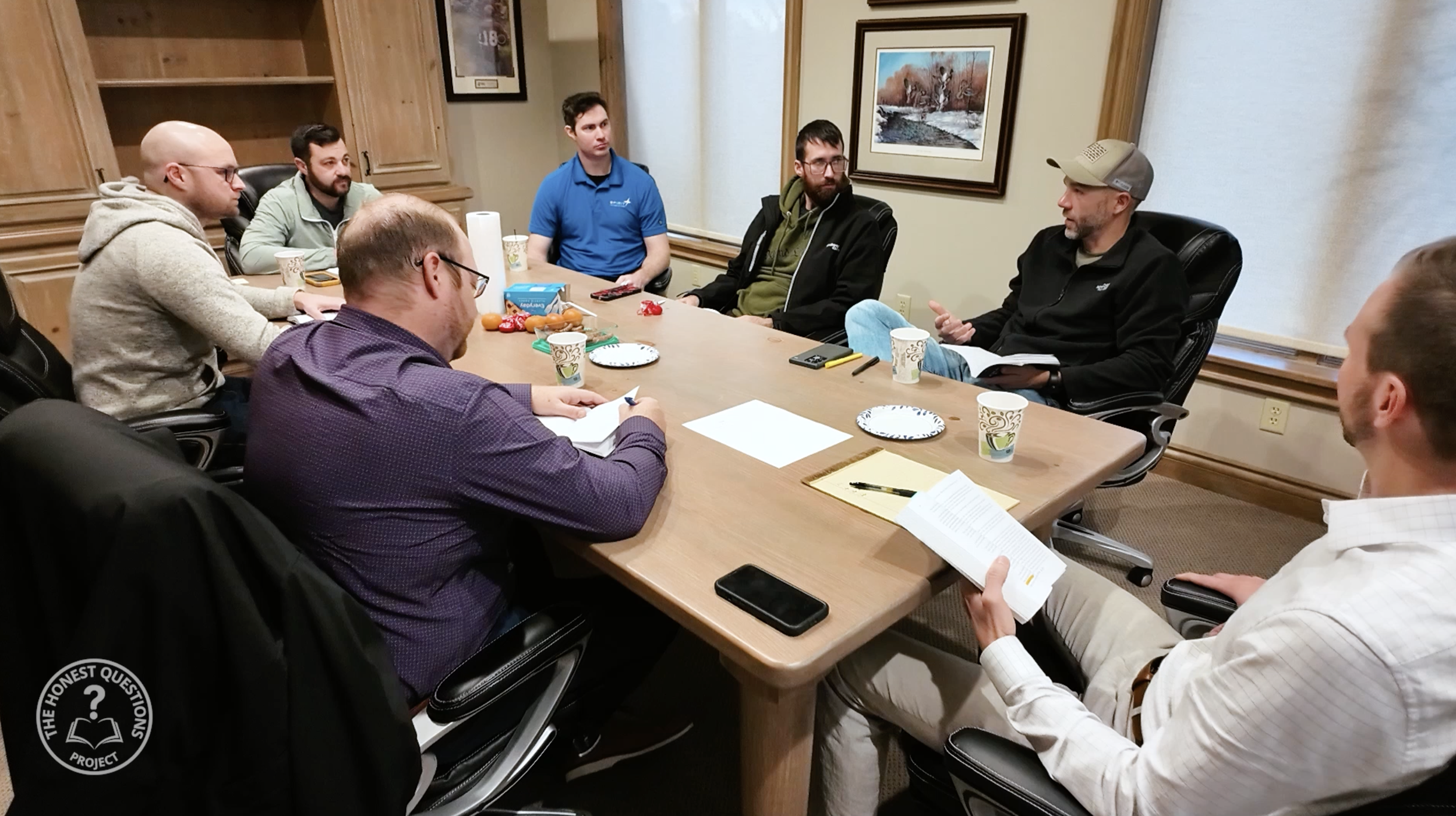 A group of eight men sitting around a conference table in a meeting room, engaged in discussion. The table has papers, notebooks, cups, and a plate of snacks. The room has wooden paneling and framed artwork on the walls.