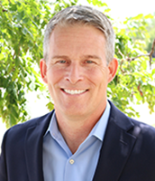 A smiling man with gray hair wearing a blue suit jacket and light blue shirt, standing outdoors with green foliage in the background.