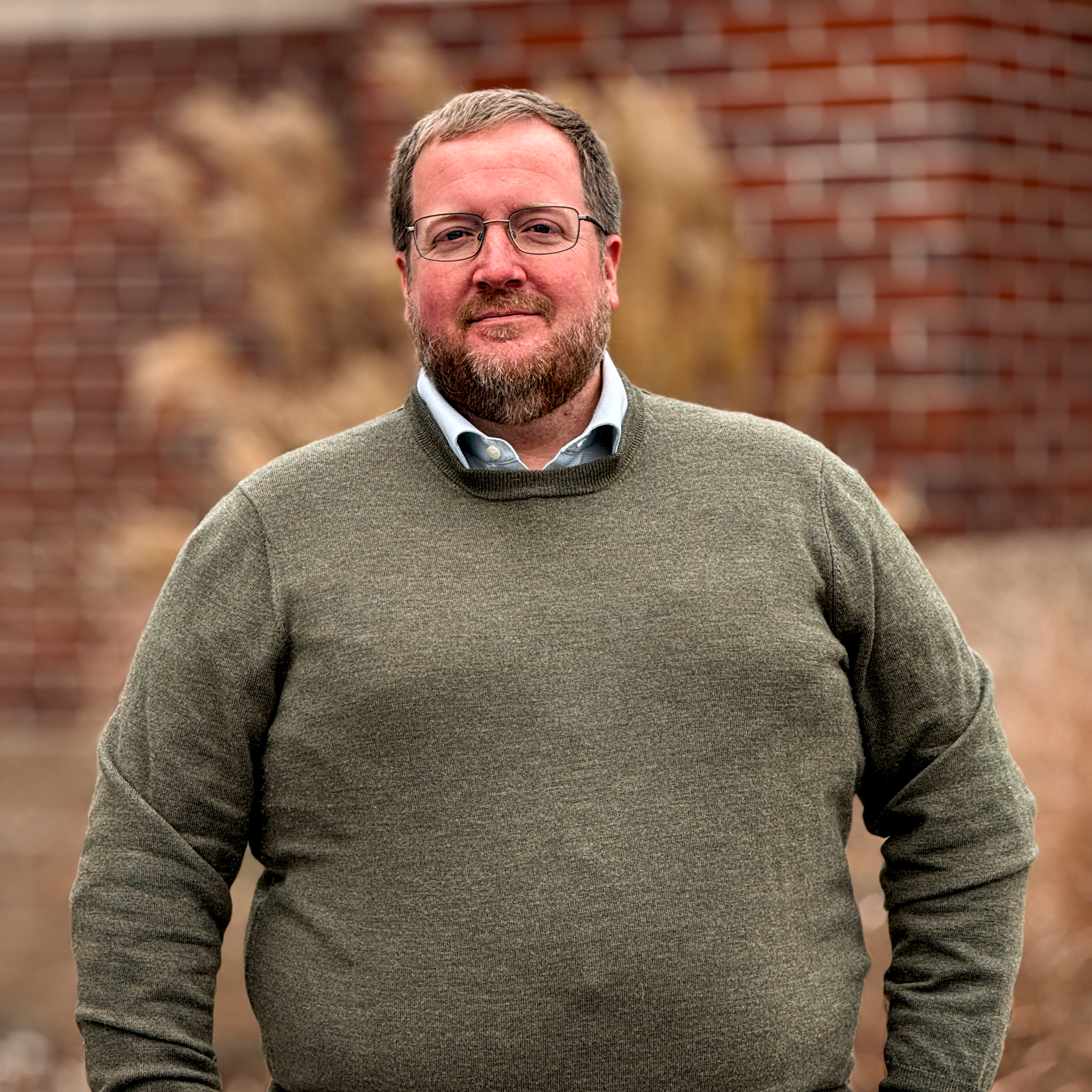 A man with glasses and a beard wearing a green sweater over a collared shirt, standing outdoors in front of a brick wall and blurred autumn foliage.