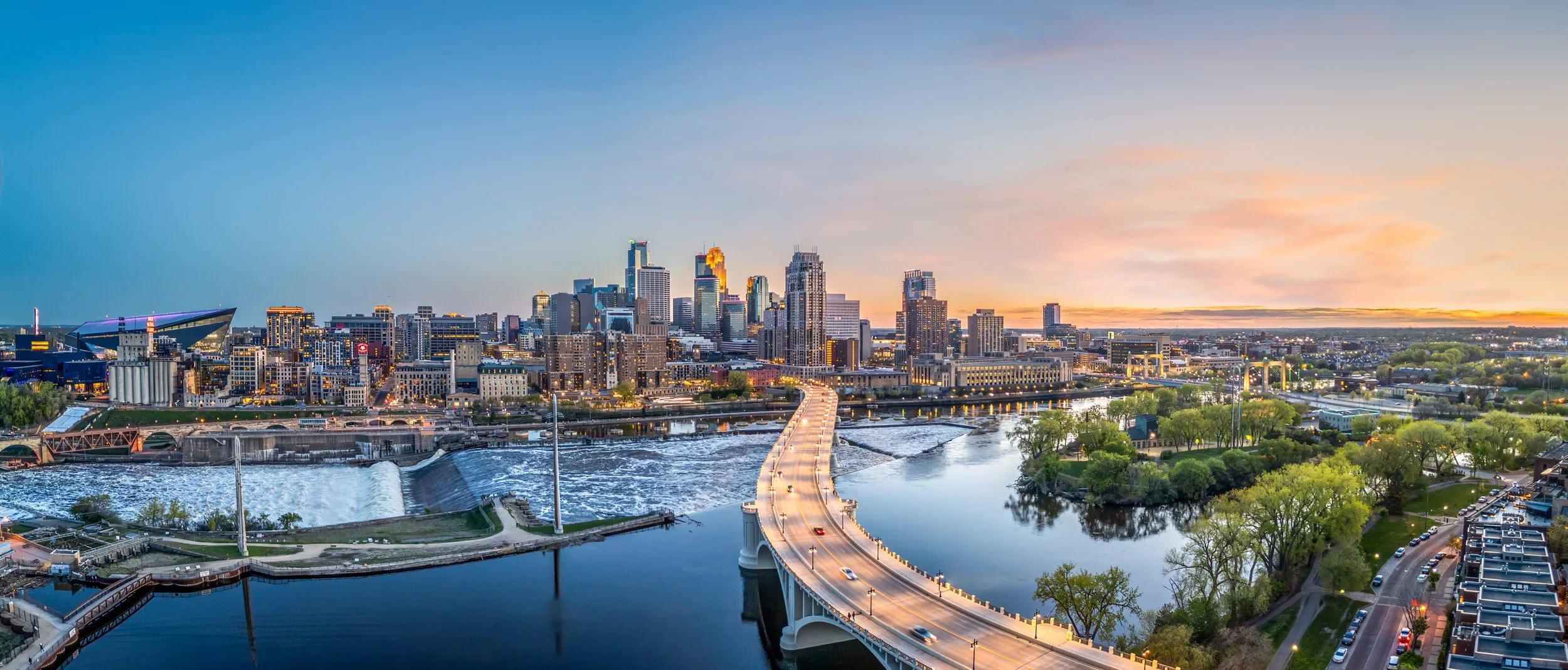 Skyline of a city at sunset with a bridge crossing a river and tall buildings in the background, city lights beginning to turn on.