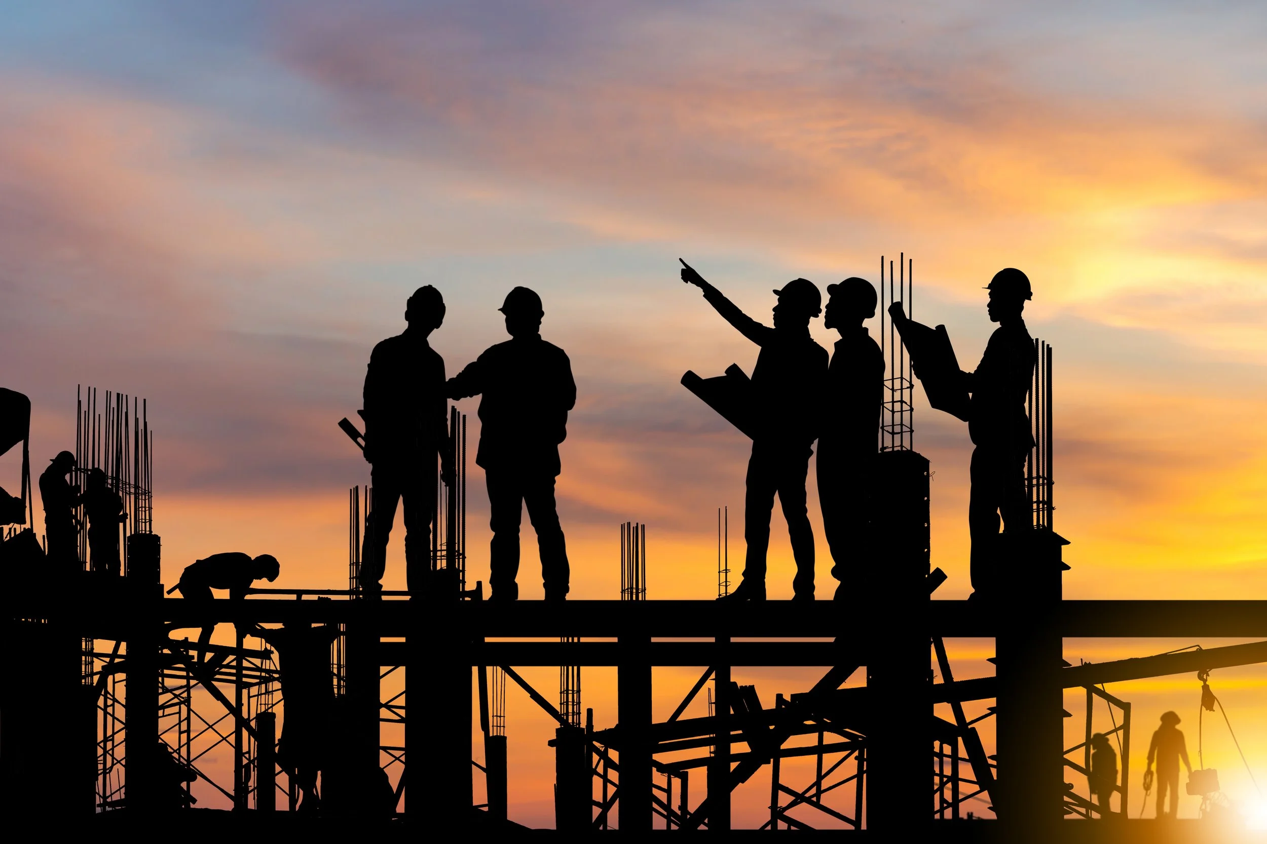 Silhouettes of construction workers on a building site during sunset, including people holding tools and discussing, with some workers on different levels of the structure against a colorful sky.
