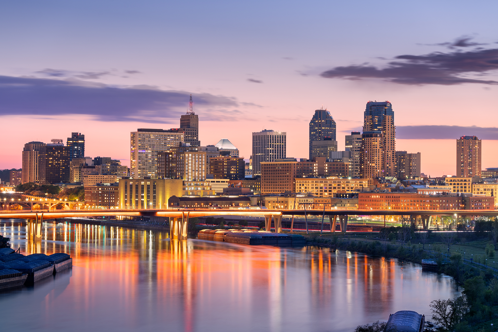 City skyline at dusk with illuminated buildings, bridge over river, and reflections on the water.