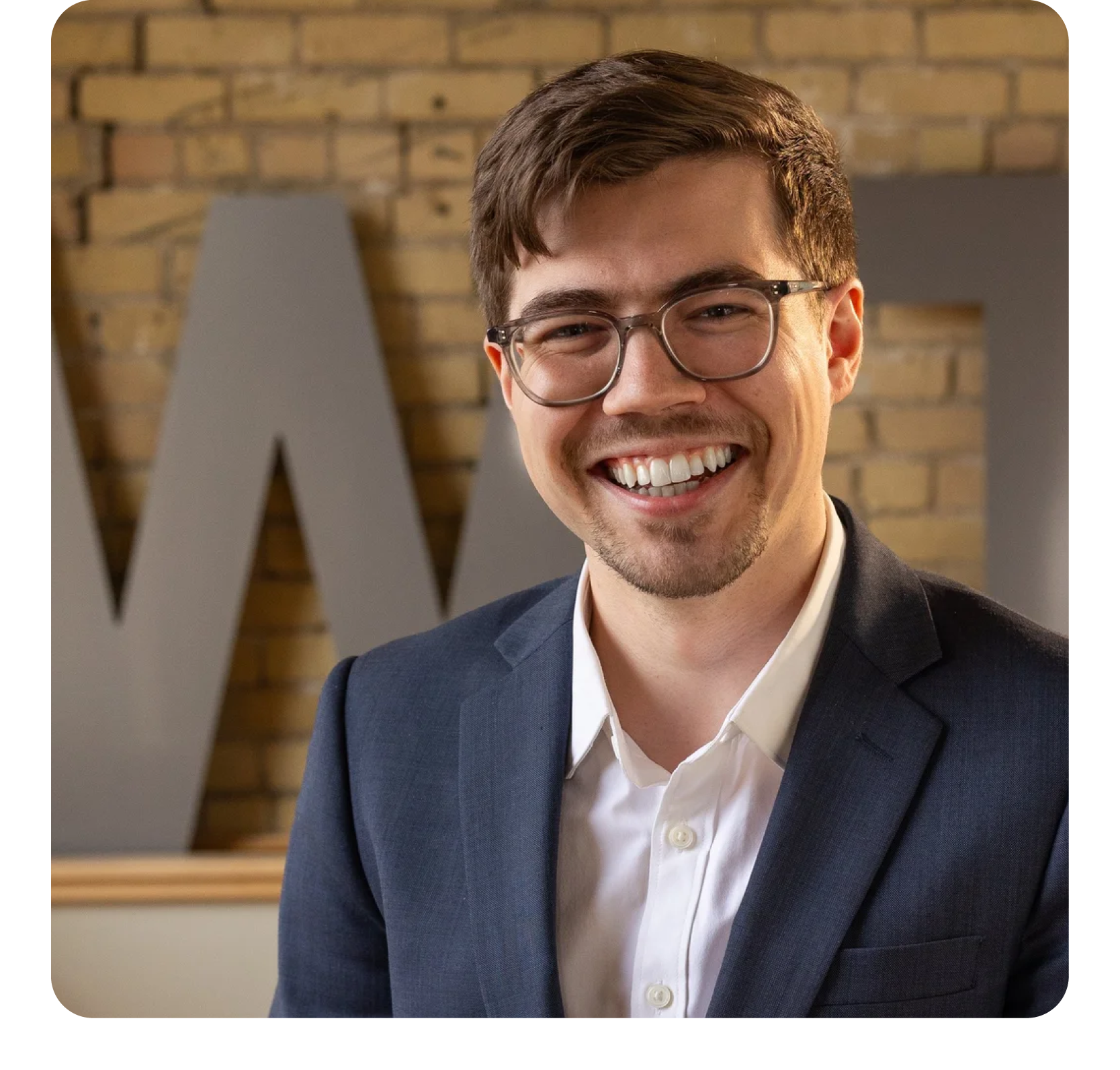 A young man with short brown hair, glasses, and a beard smiling in a professional setting. He is wearing a navy blazer and a white shirt. Behind him is a brick wall with large gray letters.