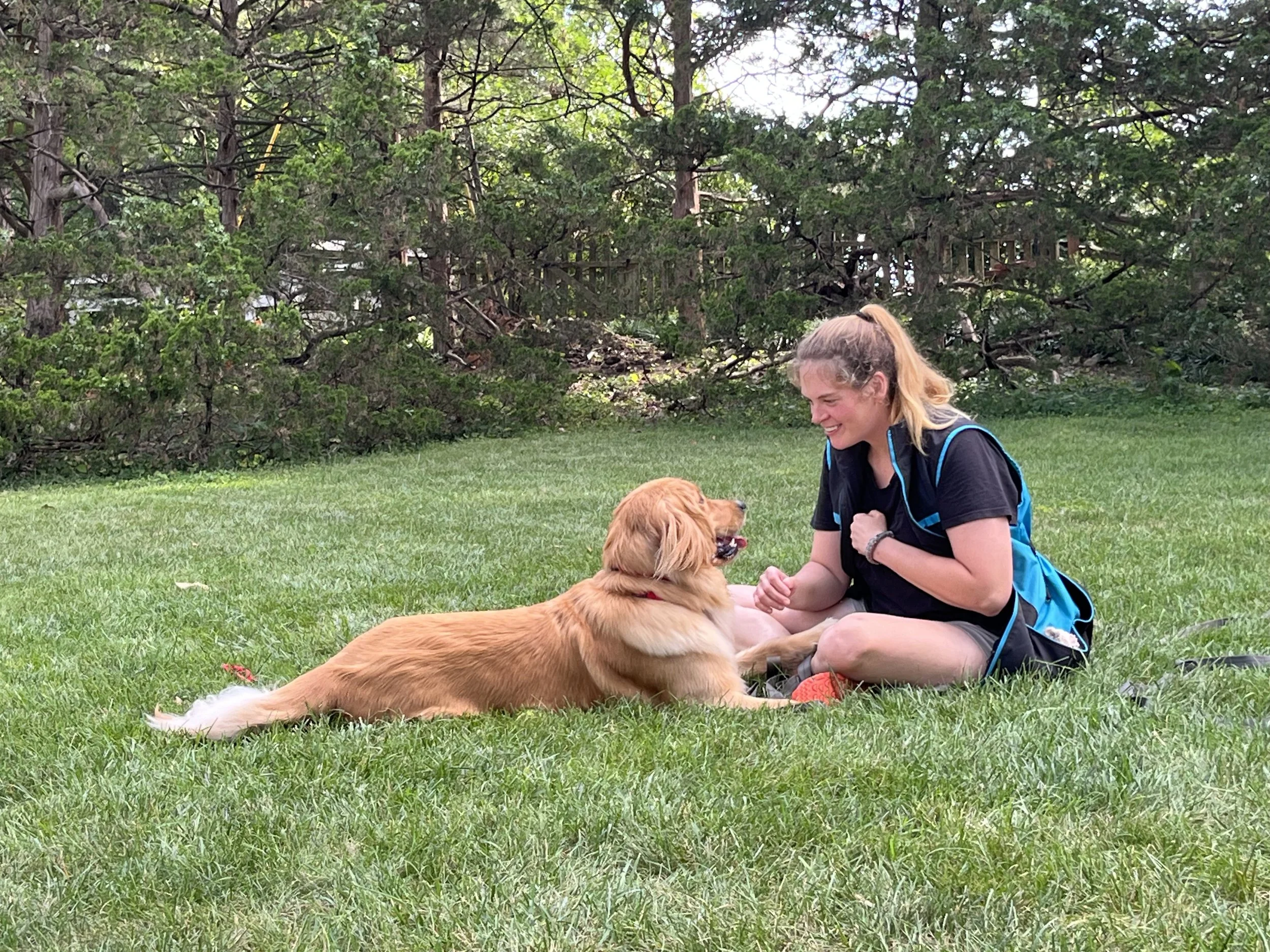 Julie Sikorak, dog trainer sitting on the grass playing with a golden retriever dog in a park with trees in the background.