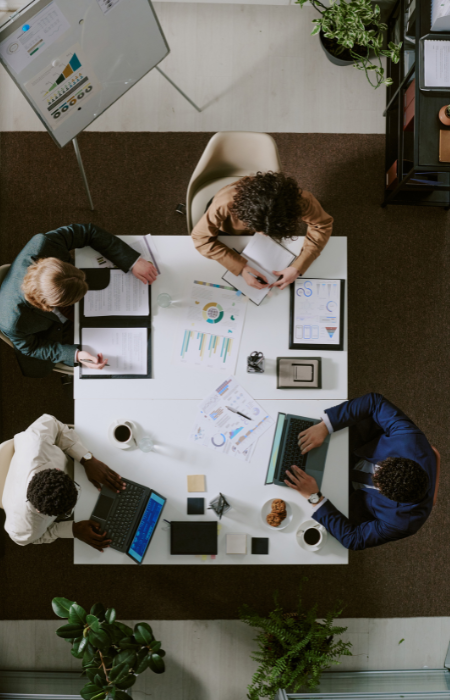 Top-down view of four people collaborating at a white office table with laptops, documents, and charts in a modern office.