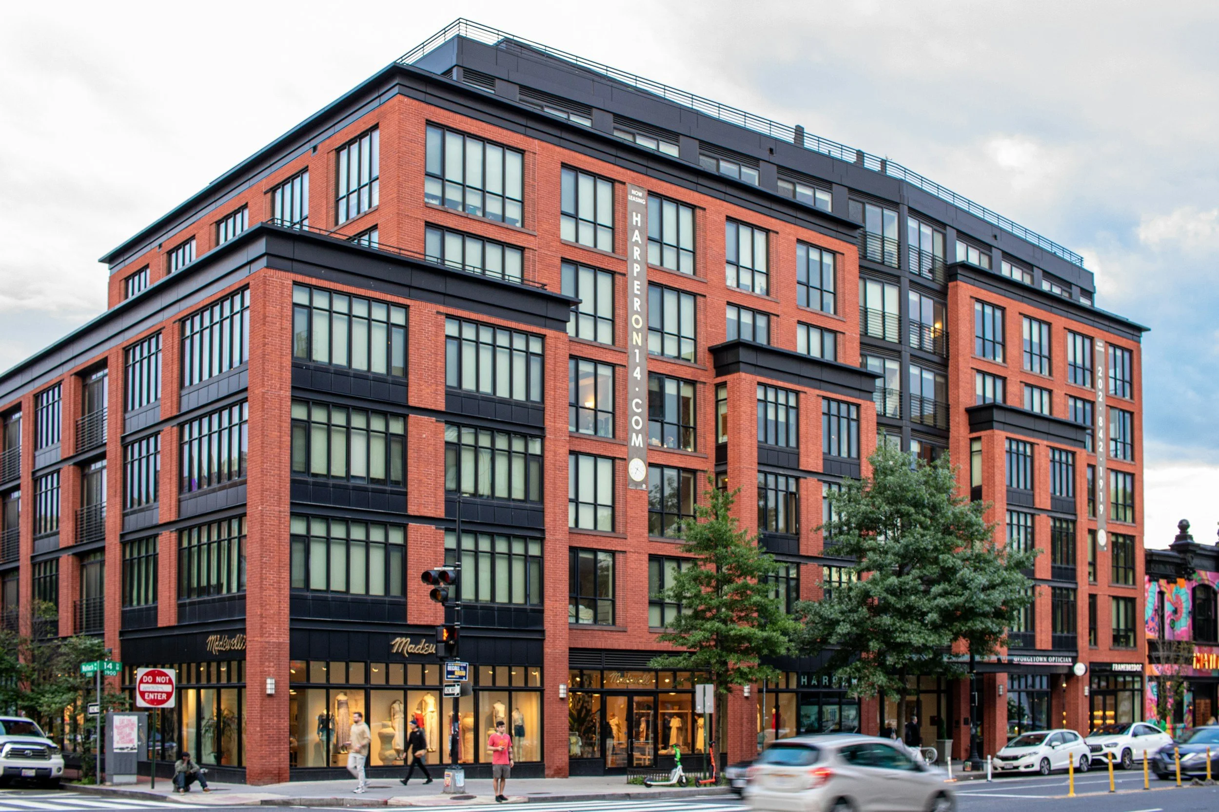 A modern, multi-story building with red brick and black accents, housing retail stores on the ground floor and apartments or offices above. There are several people walking on the sidewalk, cars parked and driving on the street, and some trees and street signs visible in the scene.