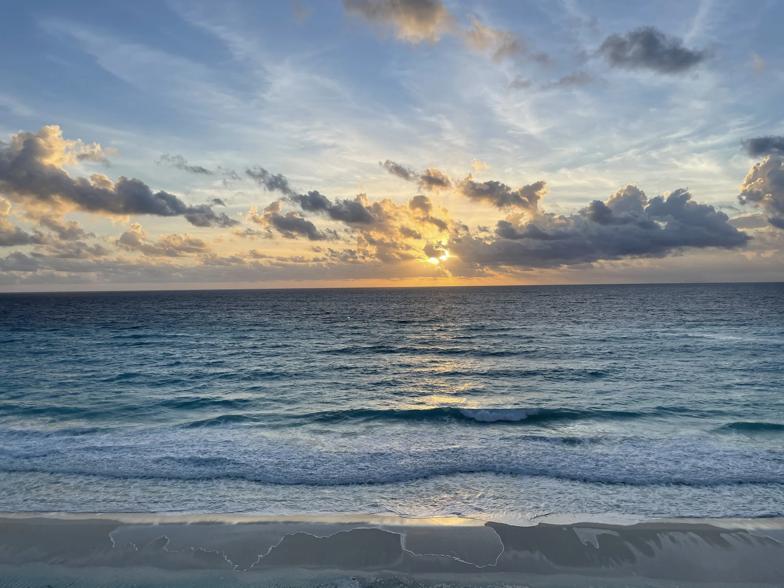 Sunset over the ocean in Cancun with clouds in the sky and gentle waves hitting the sandy shore.