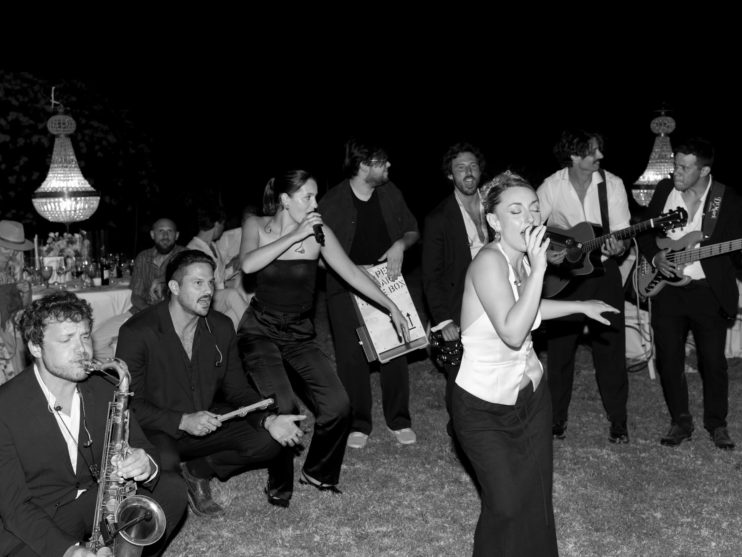 A black-and-white photo of a live music performance at night, featuring a singer with closed eyes, a band with guitars, a saxophonist, and a female vocalist holding a microphone, with elegant chandeliers and a table of guests in the background.