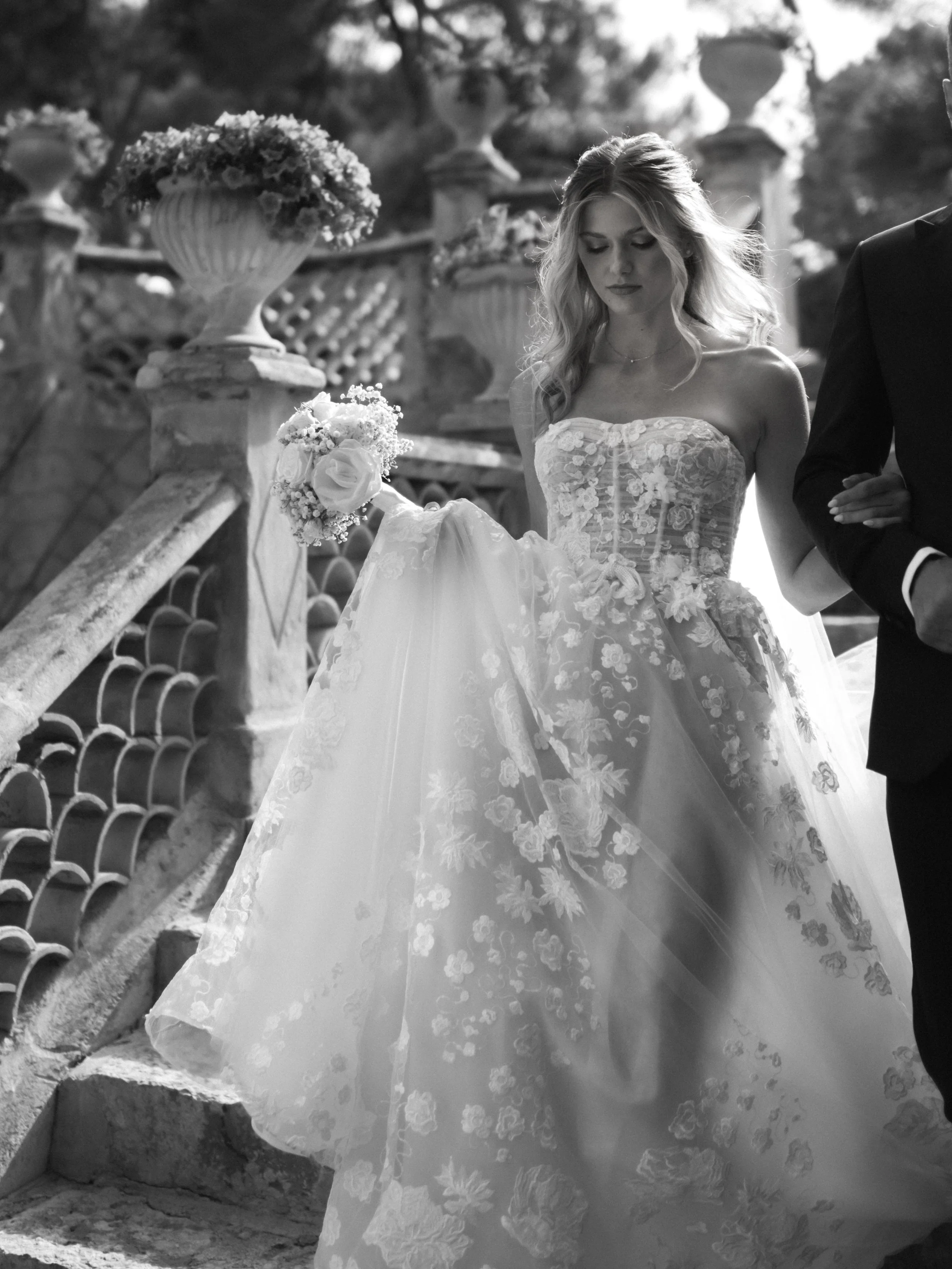A bride in a strapless floral lace wedding gown holding a bouquet on stone steps outdoors, with ornate stone balustrades and urn planters in the background.