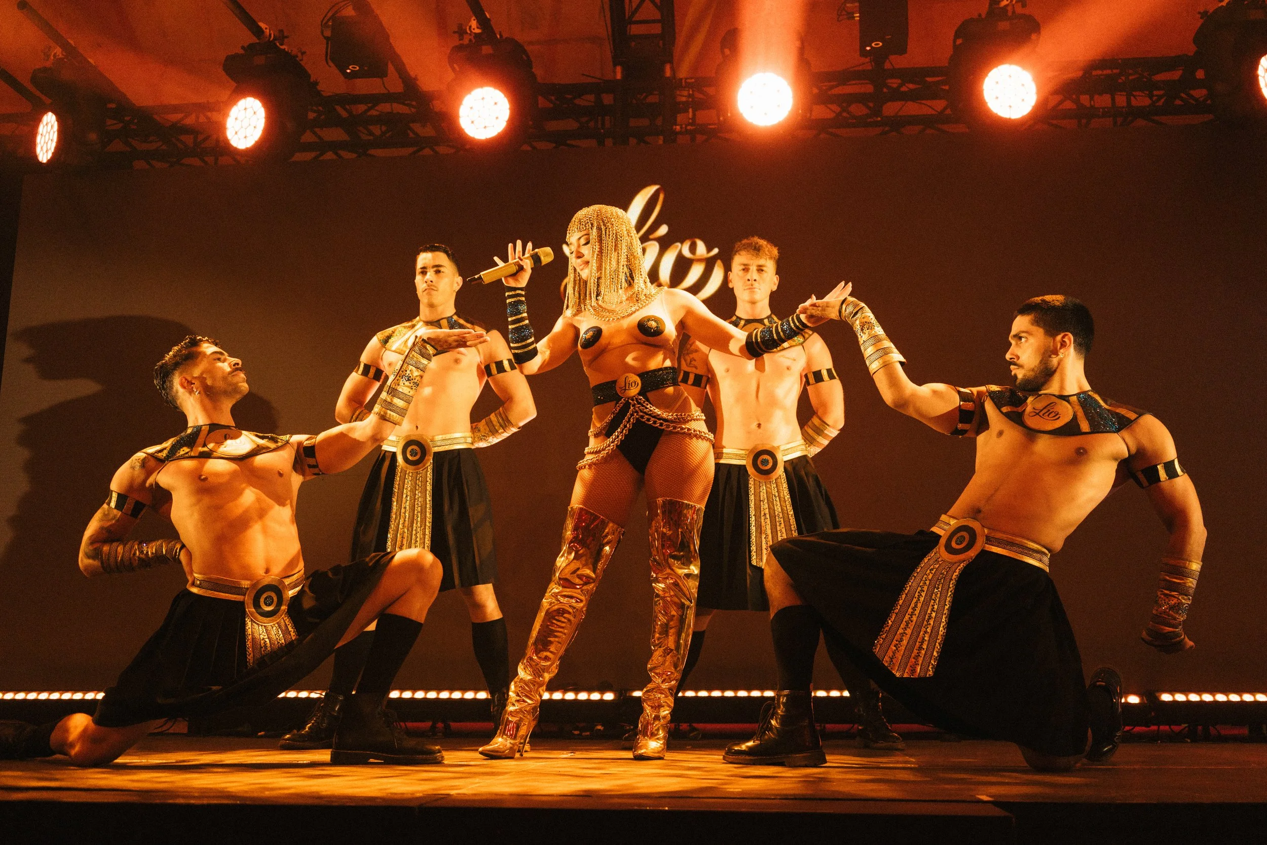 Performer with a microphone in the center, surrounded by four dancers in Egyptian-inspired costumes on stage with orange lighting.