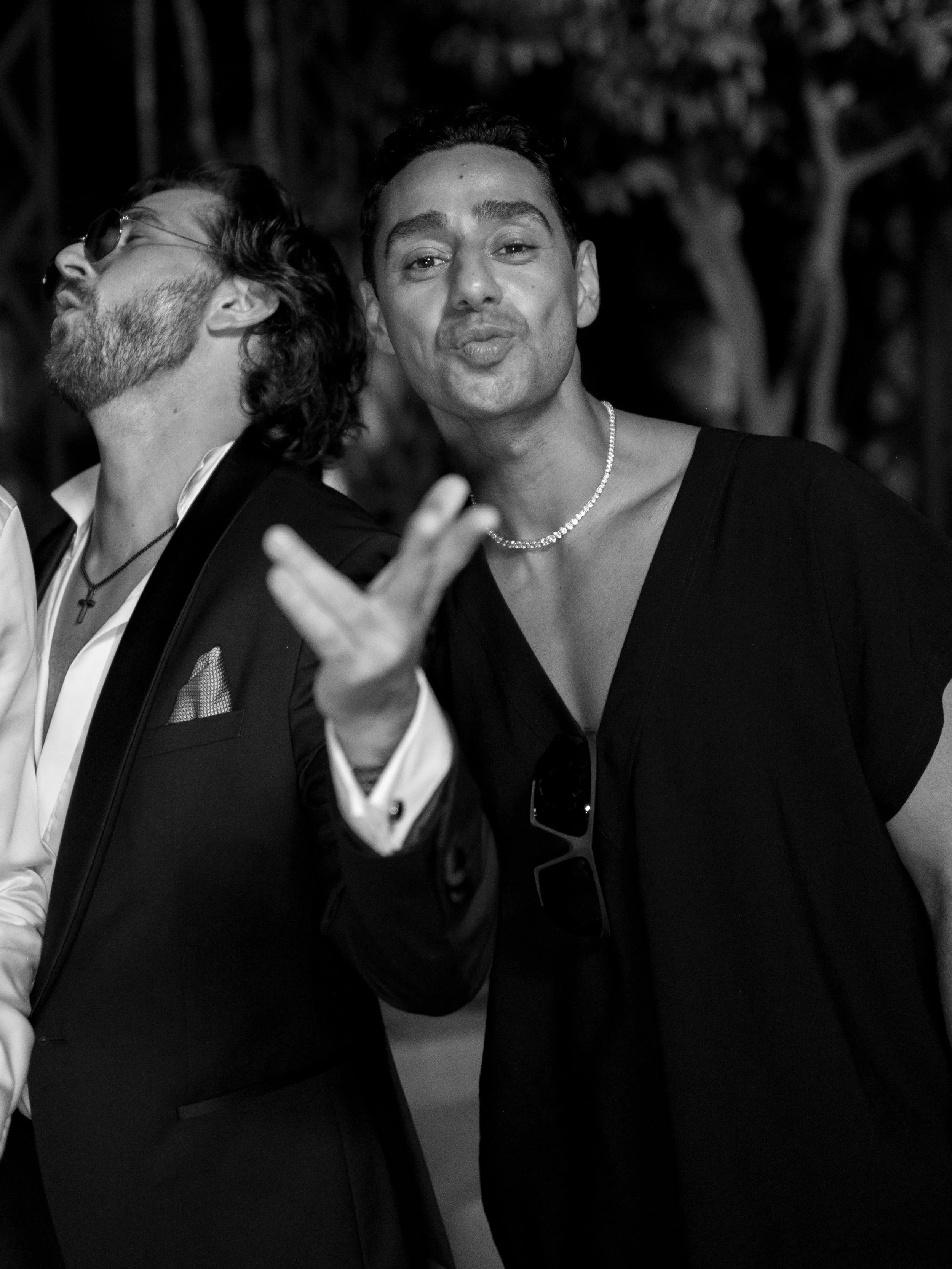 Black and white photo of two people, one with long hair and sunglasses, and one with short hair and a necklace, making a peace sign at an outdoor event.