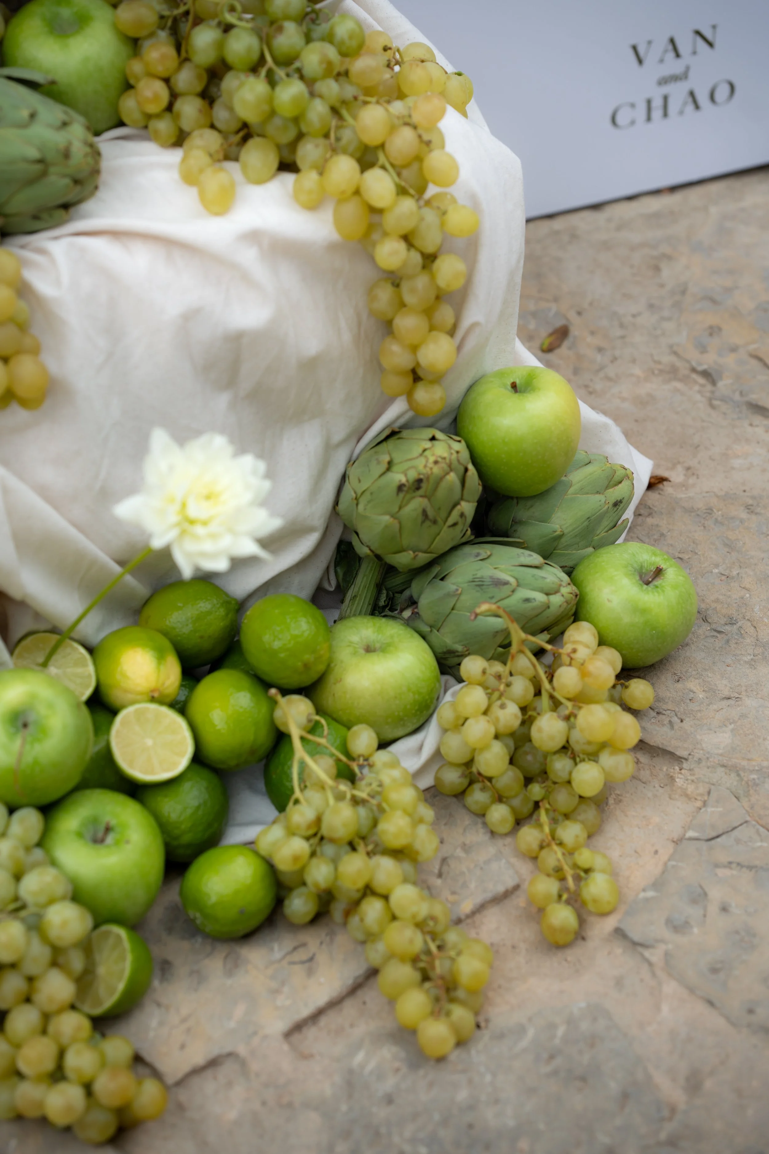 Arrangement of green apples, limes, artichokes, grapes, a lemon slice, and a white flower on a stone surface with a white cloth.