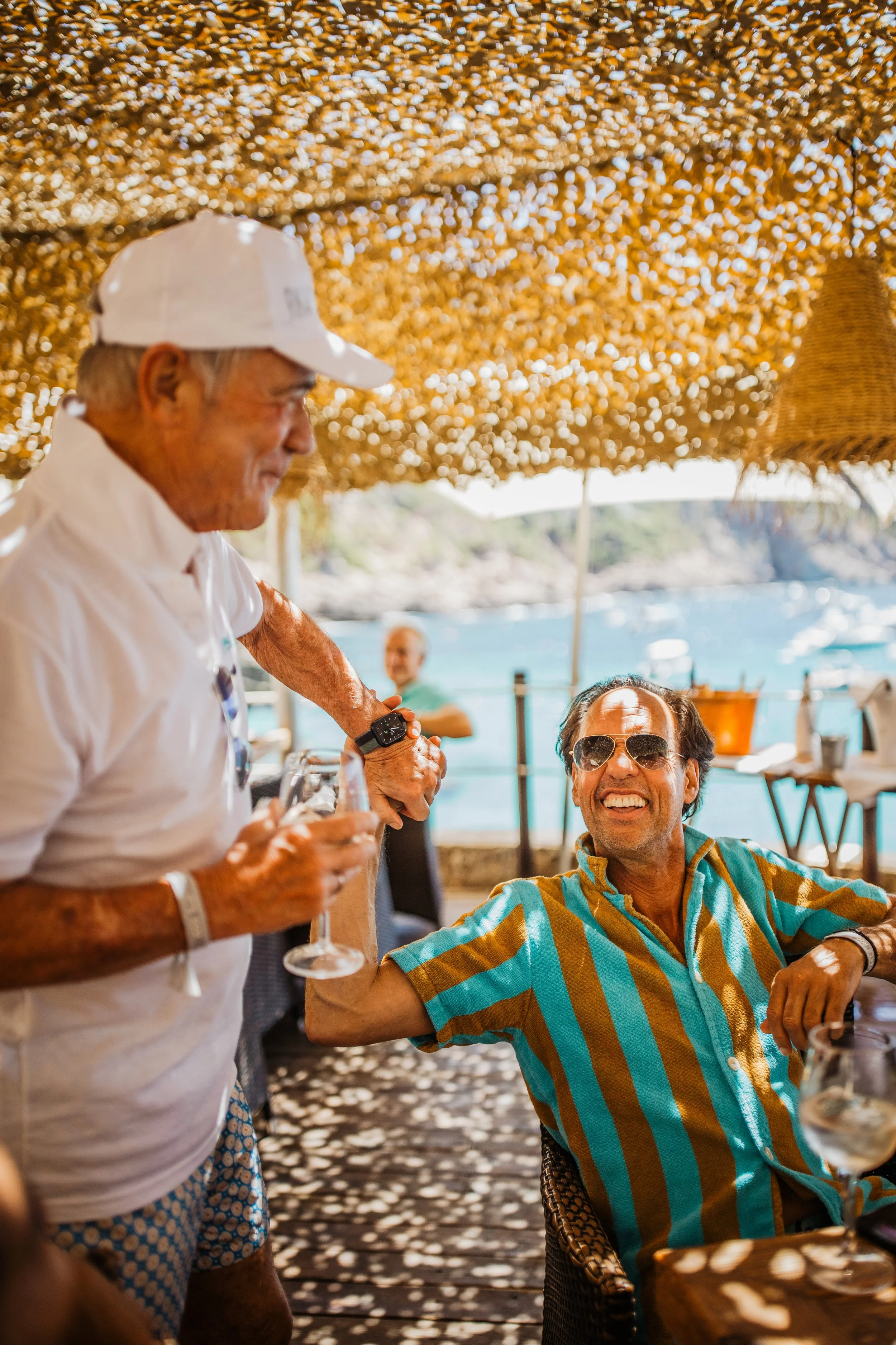 Two men enjoying drinks at an outdoor seaside restaurant under a shaded canopy, with water and a coastline visible in the background. One man is standing and holding a glass, while the other man is sitting and smiling, wearing sunglasses and a striped shirt.