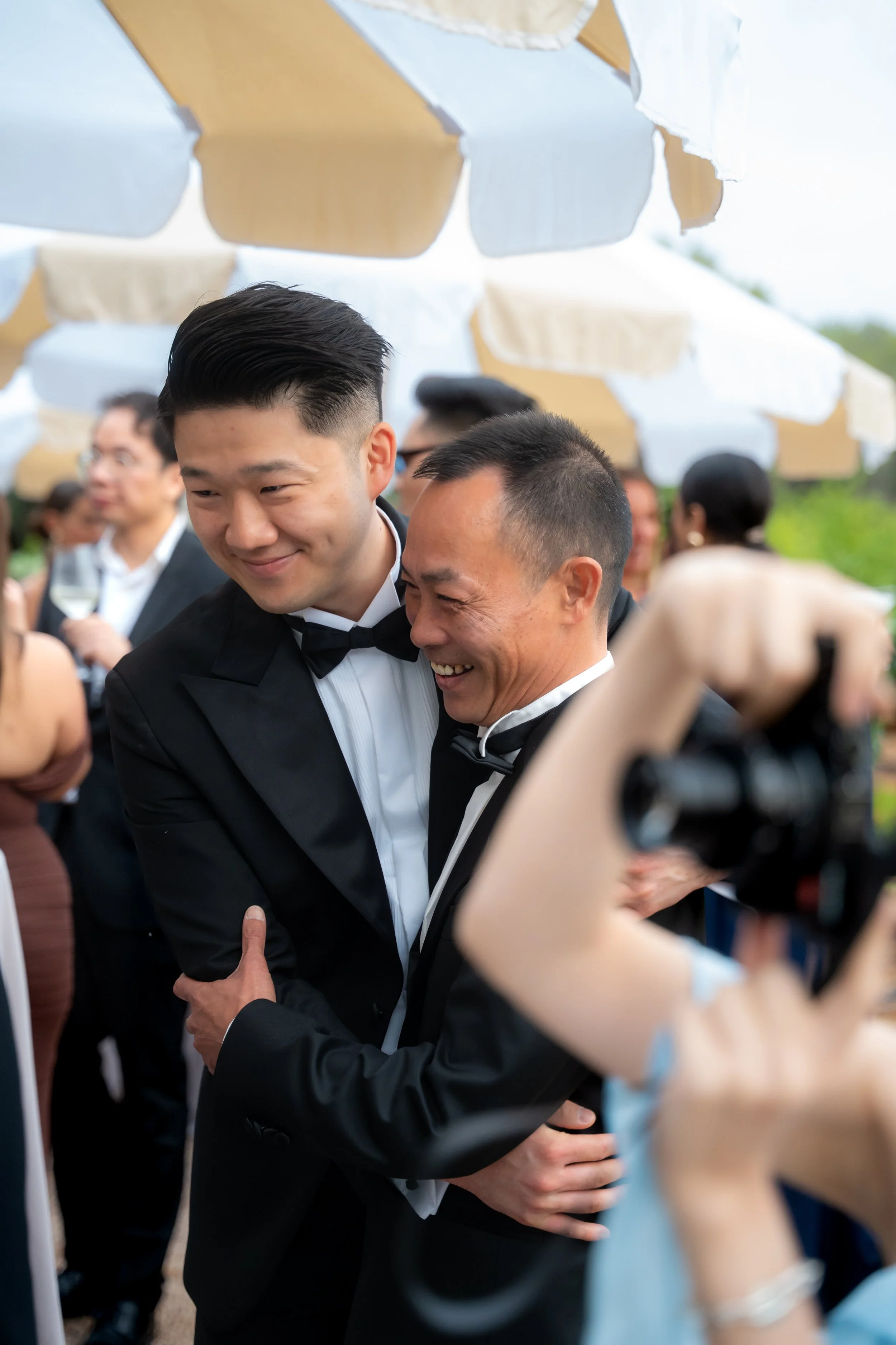Two men in tuxedos sharing a warm embrace at a social event, with other guests and umbrellas in the background.