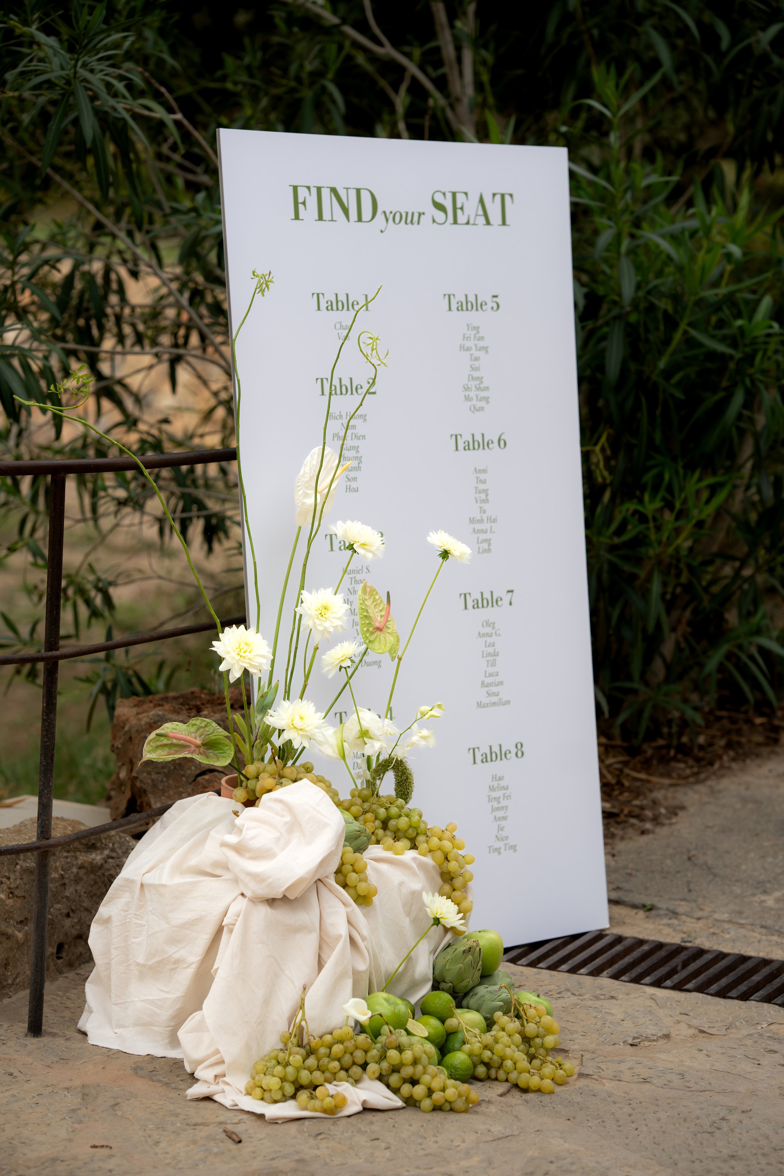 A seating chart standing behind a floral arrangement with white daisies, green grapes, and artichokes, at an outdoor event.