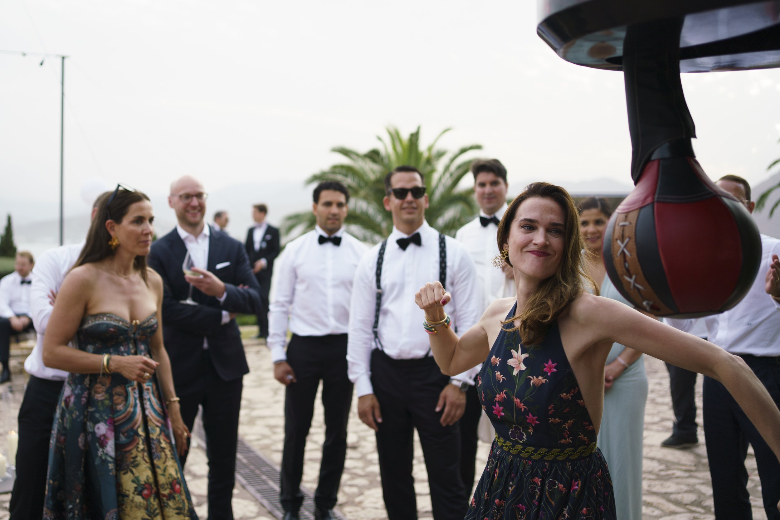 A woman in a floral dress is playing a traditional Italian game where she hits a punching bag shaped like a colorful bagpipe at an outdoor gathering. Several guests in formal attire watch and smile, with some holding drinks, as they stand on a stone 