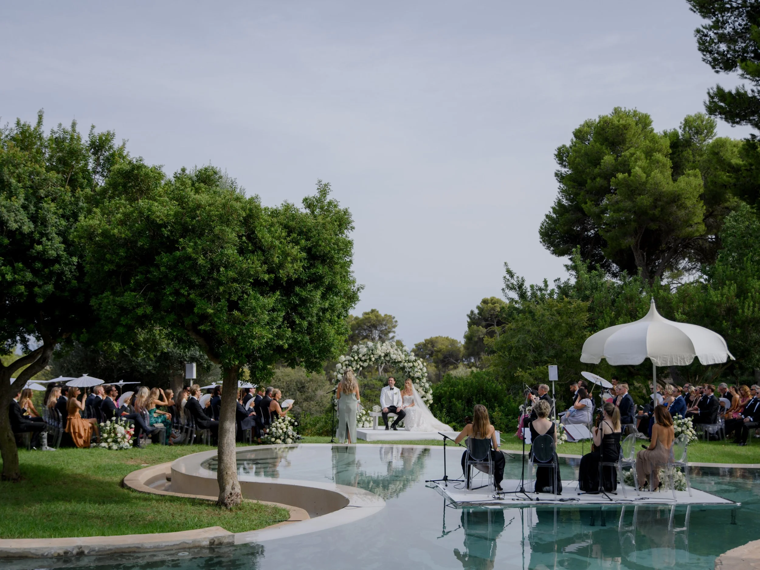 Outdoor wedding ceremony by a pool with guests seated under large trees, a bride and groom sitting at an altar with white flowers, a small band playing music nearby, and a person officiating.