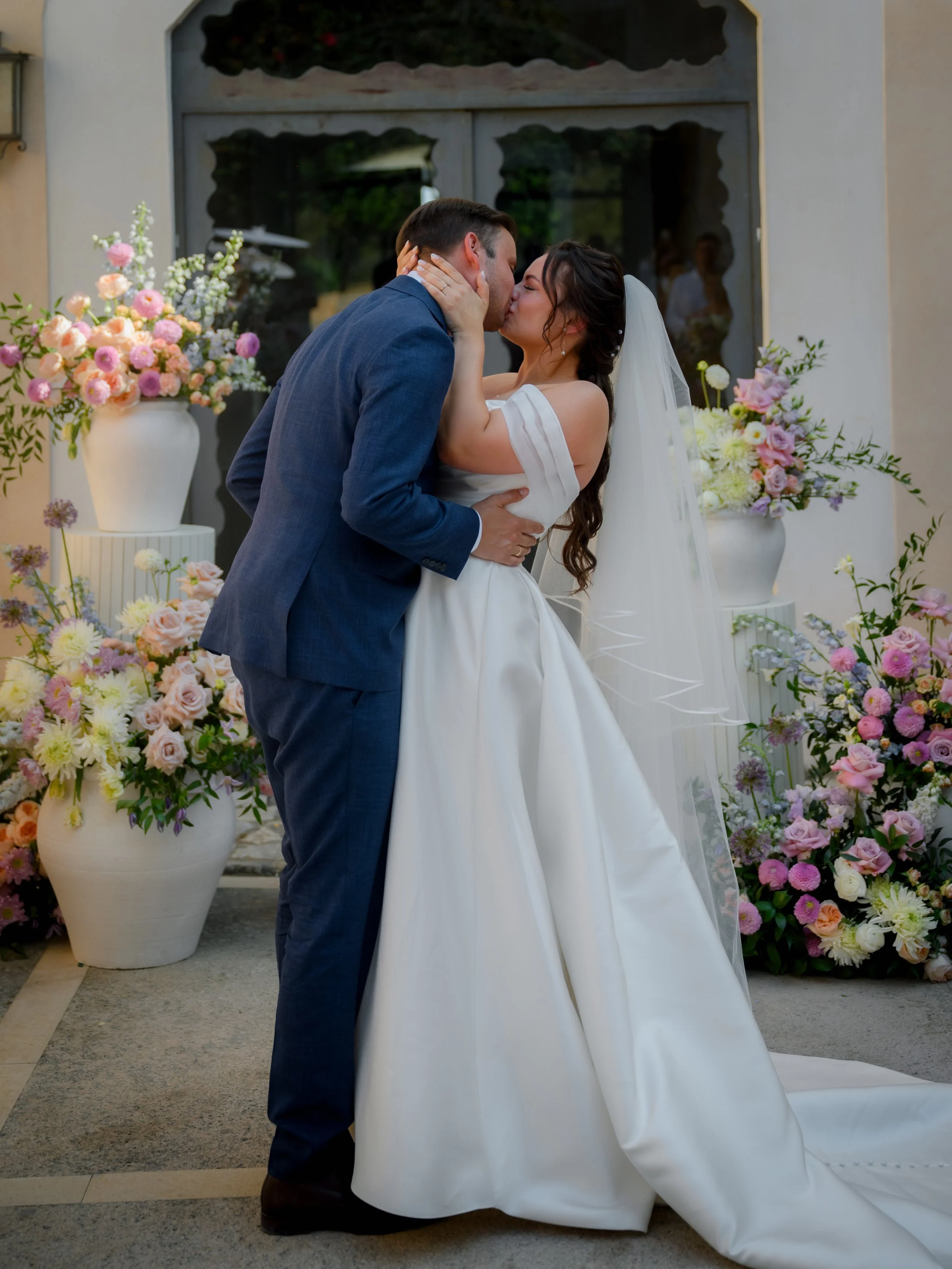 A bride and groom kiss during their wedding ceremony, surrounded by pink and white flowers in large vases.