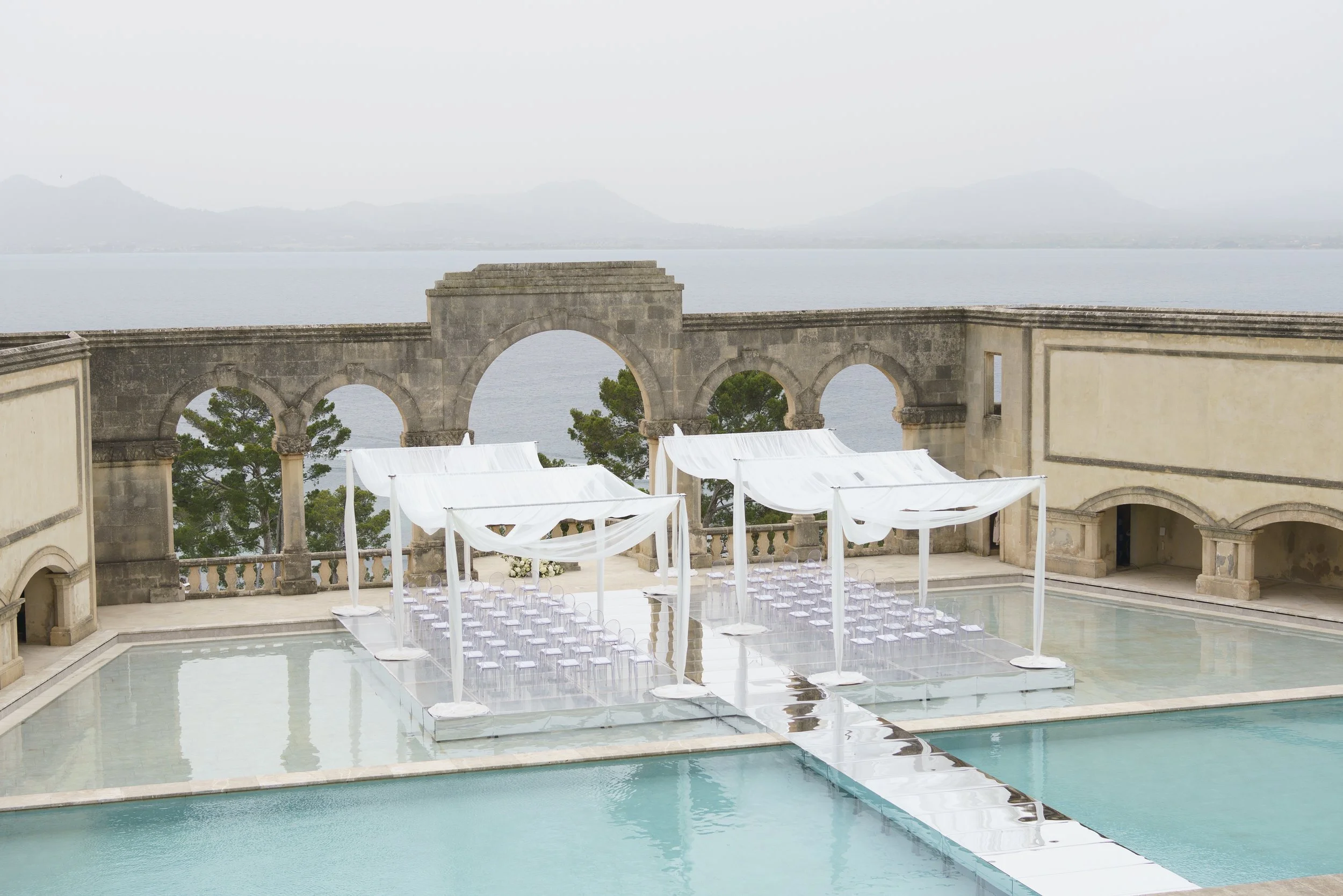 A wedding ceremony setup on a boat deck with a pool, white drapes, and chairs, overlooking a large body of water with mountains in the distance.